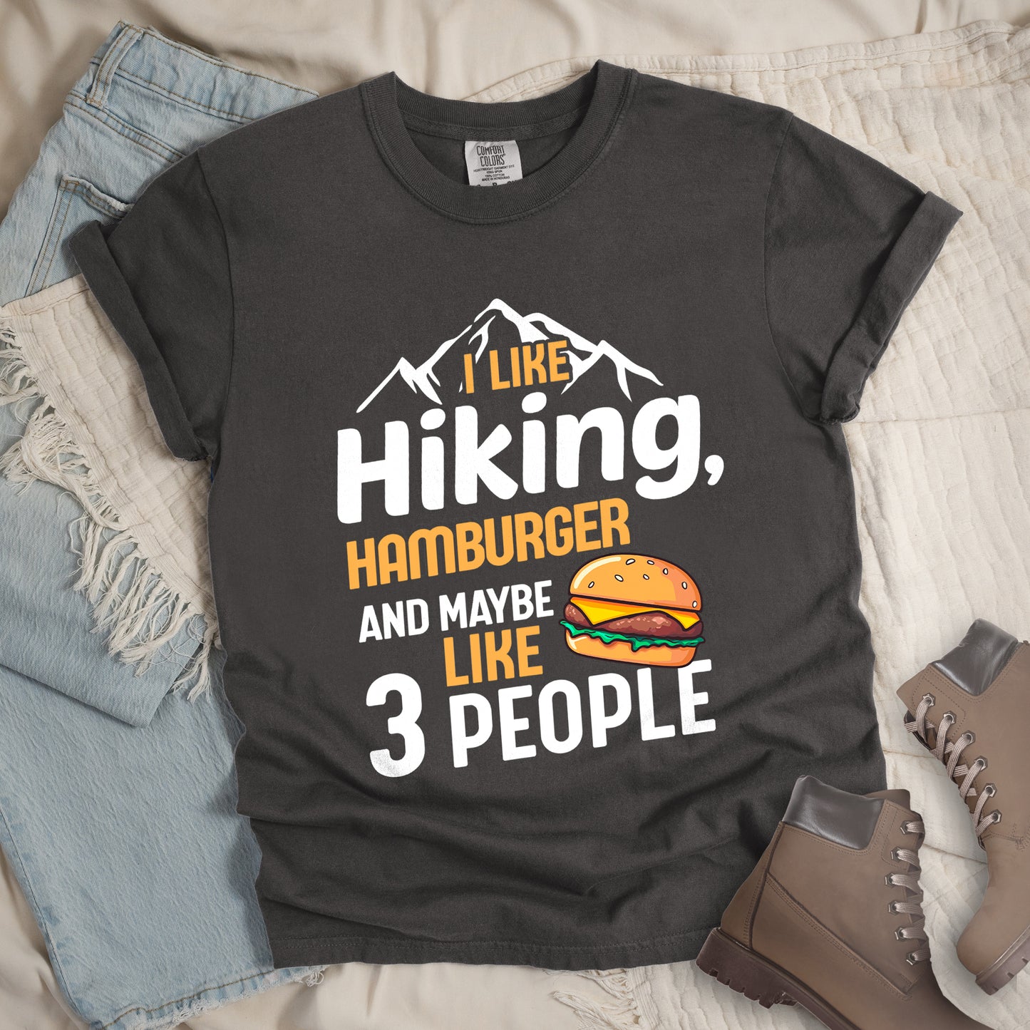 Pepper (Dark Gray) t-shirt with text "I LIKE Hiking, HAMBURGER AND MAYBE LIKE 3 PEOPLE". White mountain silhouettes appear at the top. "I LIKE" in orange, "Hiking," in white, "HAMBURGER" in orange, "AND MAYBE" in white, "LIKE" in orange, and "3 PEOPLE" in white. An illustration of a hamburger with sesame seed bun, cheese, lettuce and patty appears next to the word "LIKE". The design combines hiking and introvert humor.