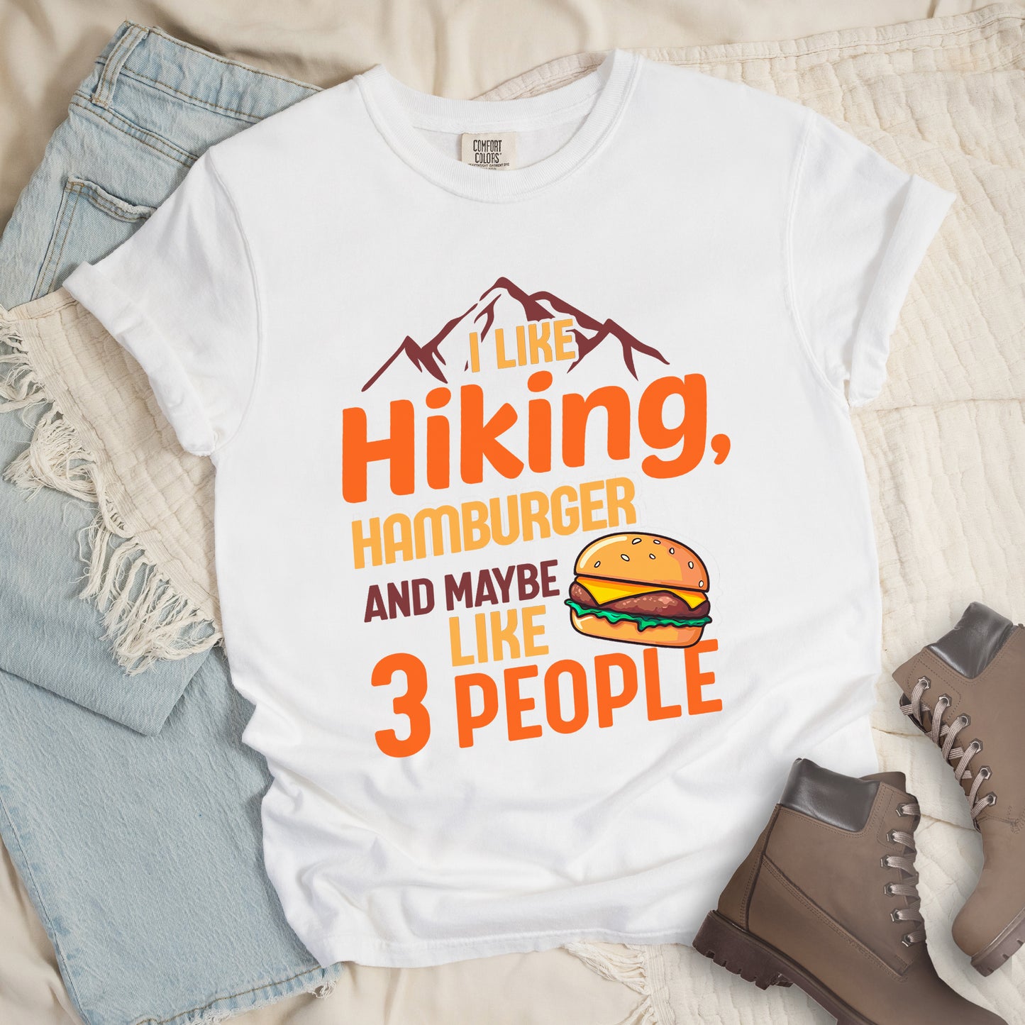 White t-shirt with text "I LIKE Hiking, HAMBURGER AND MAYBE LIKE 3 PEOPLE". Maroon mountain silhouettes appear at the top. "I LIKE" in yellow, "Hiking," in orange, "HAMBURGER" in yellow, "AND MAYBE" in maroon, "LIKE" in yellow, and "3 PEOPLE" in orange. An illustration of a hamburger with sesame seed bun, cheese, lettuce and patty appears next to the word "LIKE". The design combines hiking and introvert humor.