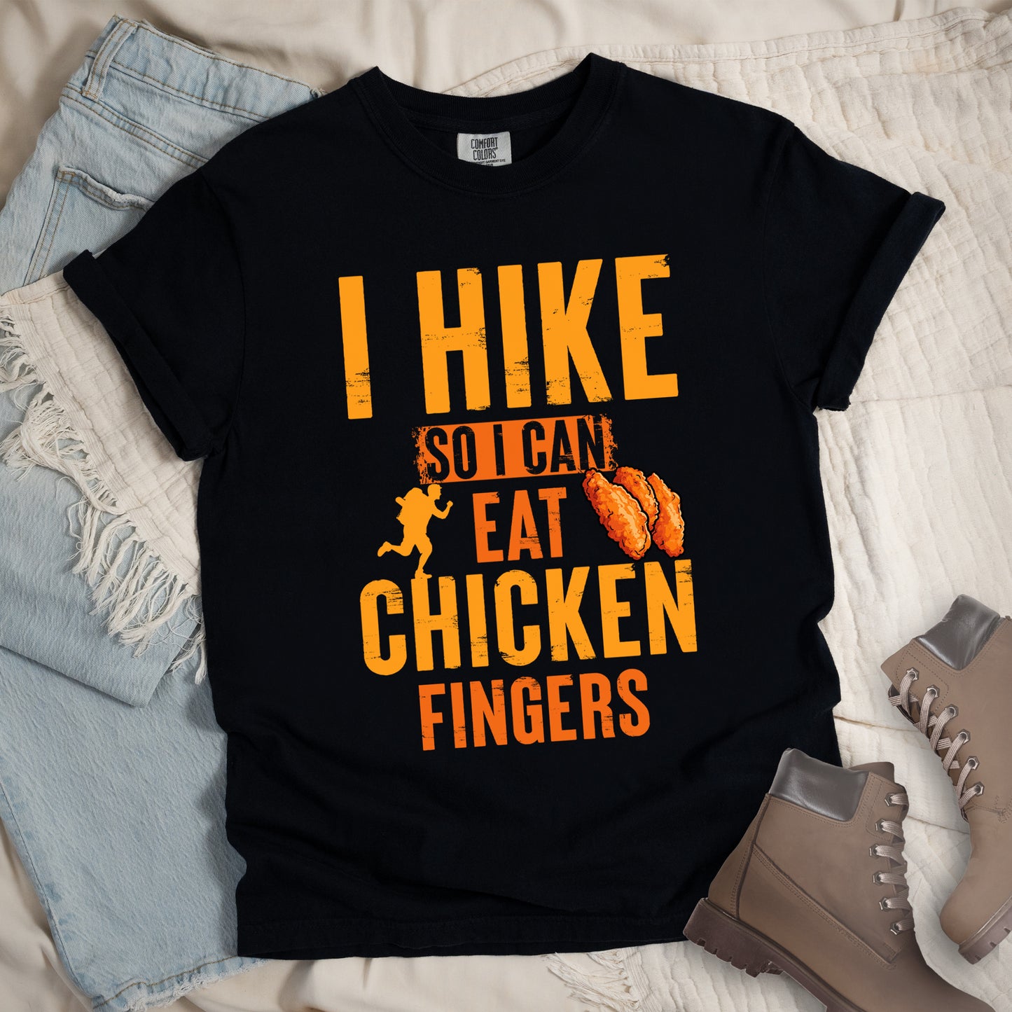 Black t-shirt with orange distressed text reading "I HIKE SO I CAN EAT CHICKEN FINGERS". All text appears in gradient orange tones from yellow-orange to deeper orange. A small hiker silhouette appears after "SO I CAN", and an illustration of three breaded chicken fingers/tenders appears next to "EAT". The design presents hiking as justification for enjoying comfort food, with a playful, casual aesthetic.