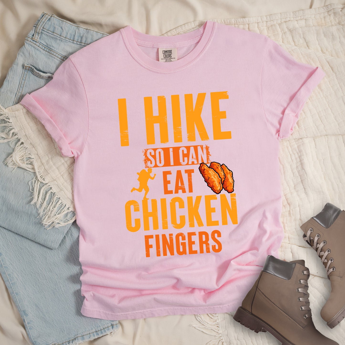 Blossom (Light Pink) t-shirt with orange distressed text reading "I HIKE SO I CAN EAT CHICKEN FINGERS". All text appears in gradient orange tones from yellow-orange to deeper orange. A small hiker silhouette appears after "SO I CAN", and an illustration of three breaded chicken fingers/tenders appears next to "EAT". The design presents hiking as justification for enjoying comfort food, with a playful, casual aesthetic.