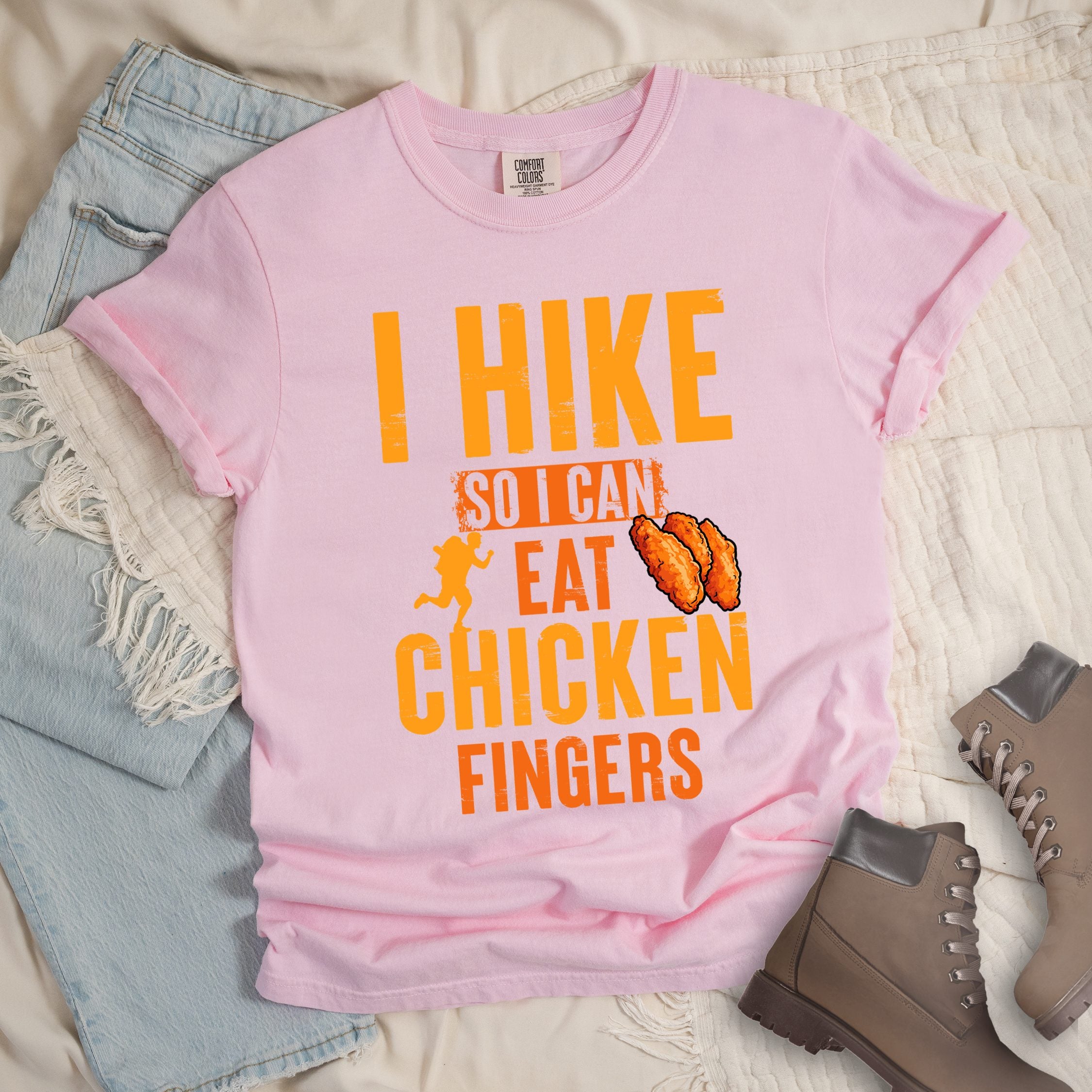 Light pink shirt with Hiking Design featuring the phrase “I Hike So I Can Eat Chicken Fingers” and an illustration of crispy chicken fingers beside a hiker silhouette.