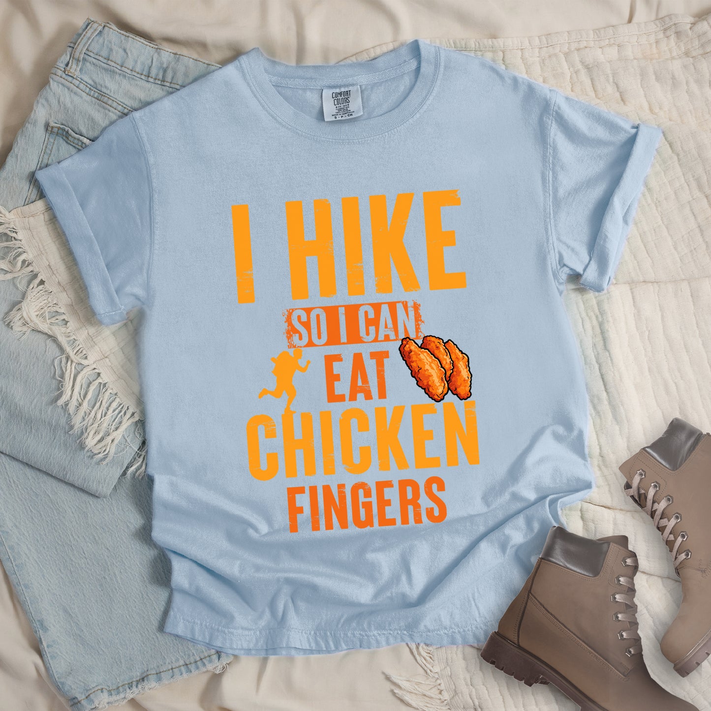 Chambray (Light Blue) t-shirt with orange distressed text reading "I HIKE SO I CAN EAT CHICKEN FINGERS". All text appears in gradient orange tones from yellow-orange to deeper orange. A small hiker silhouette appears after "SO I CAN", and an illustration of three breaded chicken fingers/tenders appears next to "EAT". The design presents hiking as justification for enjoying comfort food, with a playful, casual aesthetic.