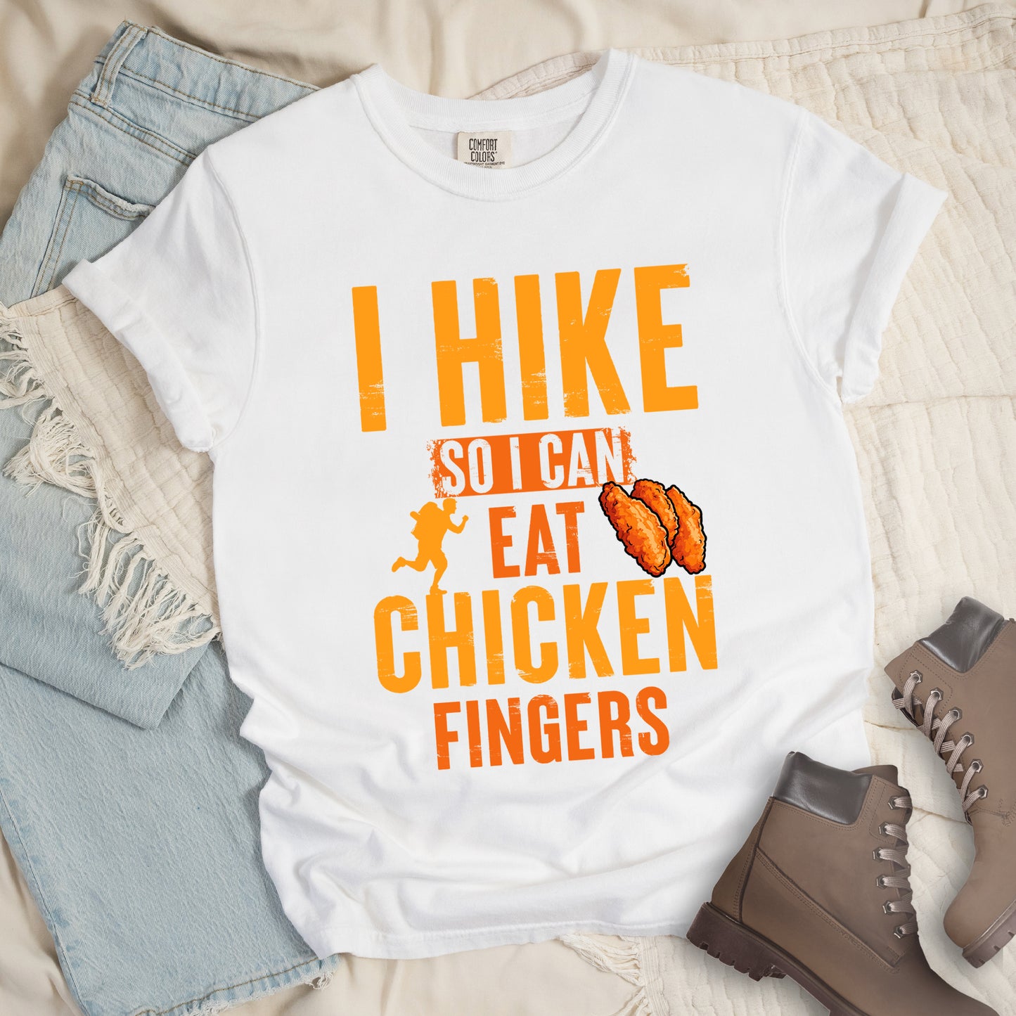 White t-shirt with orange distressed text reading "I HIKE SO I CAN EAT CHICKEN FINGERS". All text appears in gradient orange tones from yellow-orange to deeper orange. A small hiker silhouette appears after "SO I CAN", and an illustration of three breaded chicken fingers/tenders appears next to "EAT". The design presents hiking as justification for enjoying comfort food, with a playful, casual aesthetic.
