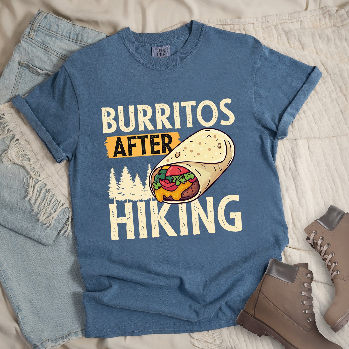 Blue Jean (Denim Blue) t-shirt with distressed text reading "BURRITOS AFTER HIKING". "BURRITOS" appears at the top in cream/off-white letters, "AFTER" in black text on an orange background in the middle, and "HIKING" in matching cream letters at the bottom. Between the text is an illustration of a burrito with visible fillings (beans, cheese, lettuce, tomato) and pine tree silhouettes in cream color. The design has a vintage, weathered appearance.