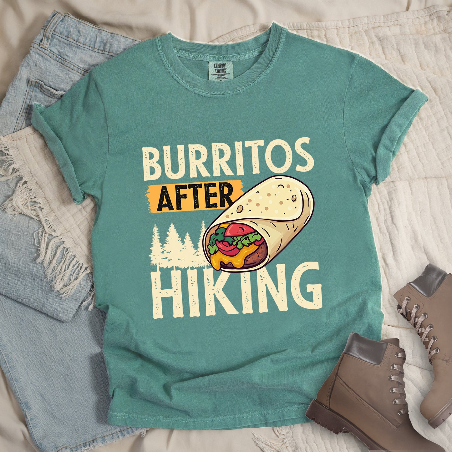 Light Green t-shirt with distressed text reading "BURRITOS AFTER HIKING". "BURRITOS" appears at the top in cream/off-white letters, "AFTER" in black text on an orange background in the middle, and "HIKING" in matching cream letters at the bottom. Between the text is an illustration of a burrito with visible fillings (beans, cheese, lettuce, tomato) and pine tree silhouettes in cream color. The design has a vintage, weathered appearance.