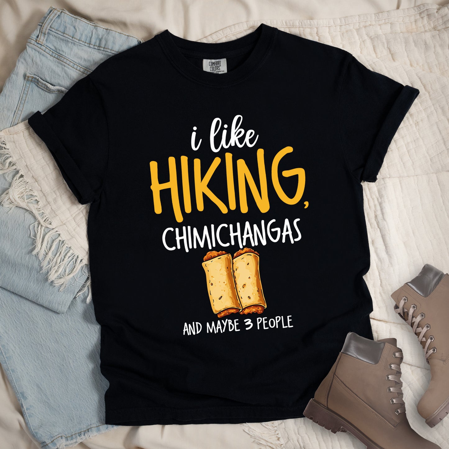 Black t-shirt with text "i like HIKING, CHIMICHANGAS AND MAYBE 3 PEOPLE". The phrase "i like" appears in white script at the top, "HIKING," in yellow letters, "CHIMICHANGAS" in white, and "AND MAYBE 3 PEOPLE" in white at the bottom. Between "CHIMICHANGAS" and "AND MAYBE 3 PEOPLE" is an illustration of two chimichangas (fried burritos) with visible fillings. The design combines hiking, food, and introvert humor.