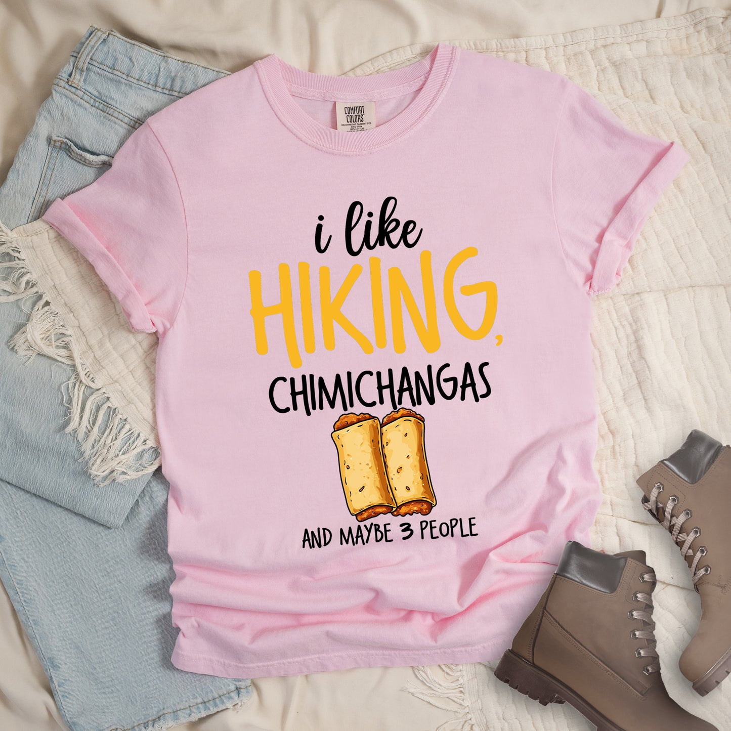 Blossom (Light Pink) t-shirt with text "i like HIKING, CHIMICHANGAS AND MAYBE 3 PEOPLE". The phrase "i like" appears in black script at the top, "HIKING," in yellow letters, "CHIMICHANGAS" in black, and "AND MAYBE 3 PEOPLE" in black at the bottom. Between "CHIMICHANGAS" and "AND MAYBE 3 PEOPLE" is an illustration of two chimichangas (fried burritos) with visible fillings. The design combines hiking, food, and introvert humor.