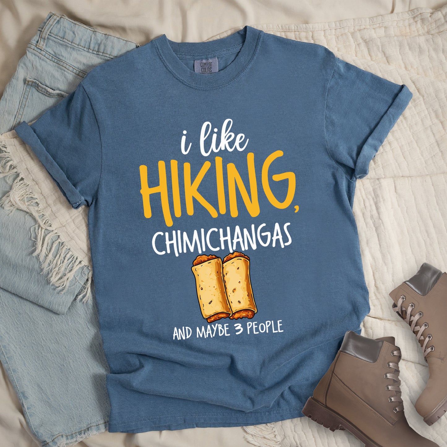 Blue Jean (Denim Blue) t-shirt with text "i like HIKING, CHIMICHANGAS AND MAYBE 3 PEOPLE". The phrase "i like" appears in white script at the top, "HIKING," in yellow letters, "CHIMICHANGAS" in white, and "AND MAYBE 3 PEOPLE" in white at the bottom. Between "CHIMICHANGAS" and "AND MAYBE 3 PEOPLE" is an illustration of two chimichangas (fried burritos) with visible fillings. The design combines hiking, food, and introvert humor.