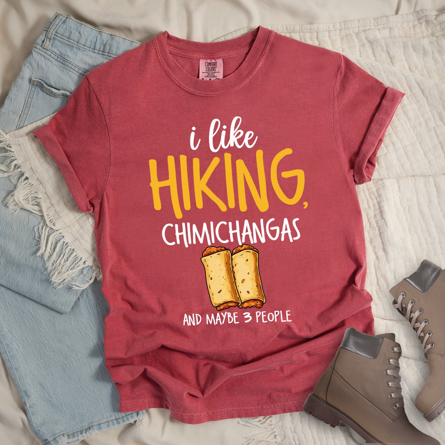 Crimson (Dark Pink) t-shirt with text "i like HIKING, CHIMICHANGAS AND MAYBE 3 PEOPLE". The phrase "i like" appears in white script at the top, "HIKING," in yellow letters, "CHIMICHANGAS" in white, and "AND MAYBE 3 PEOPLE" in white at the bottom. Between "CHIMICHANGAS" and "AND MAYBE 3 PEOPLE" is an illustration of two chimichangas (fried burritos) with visible fillings. The design combines hiking, food, and introvert humor.