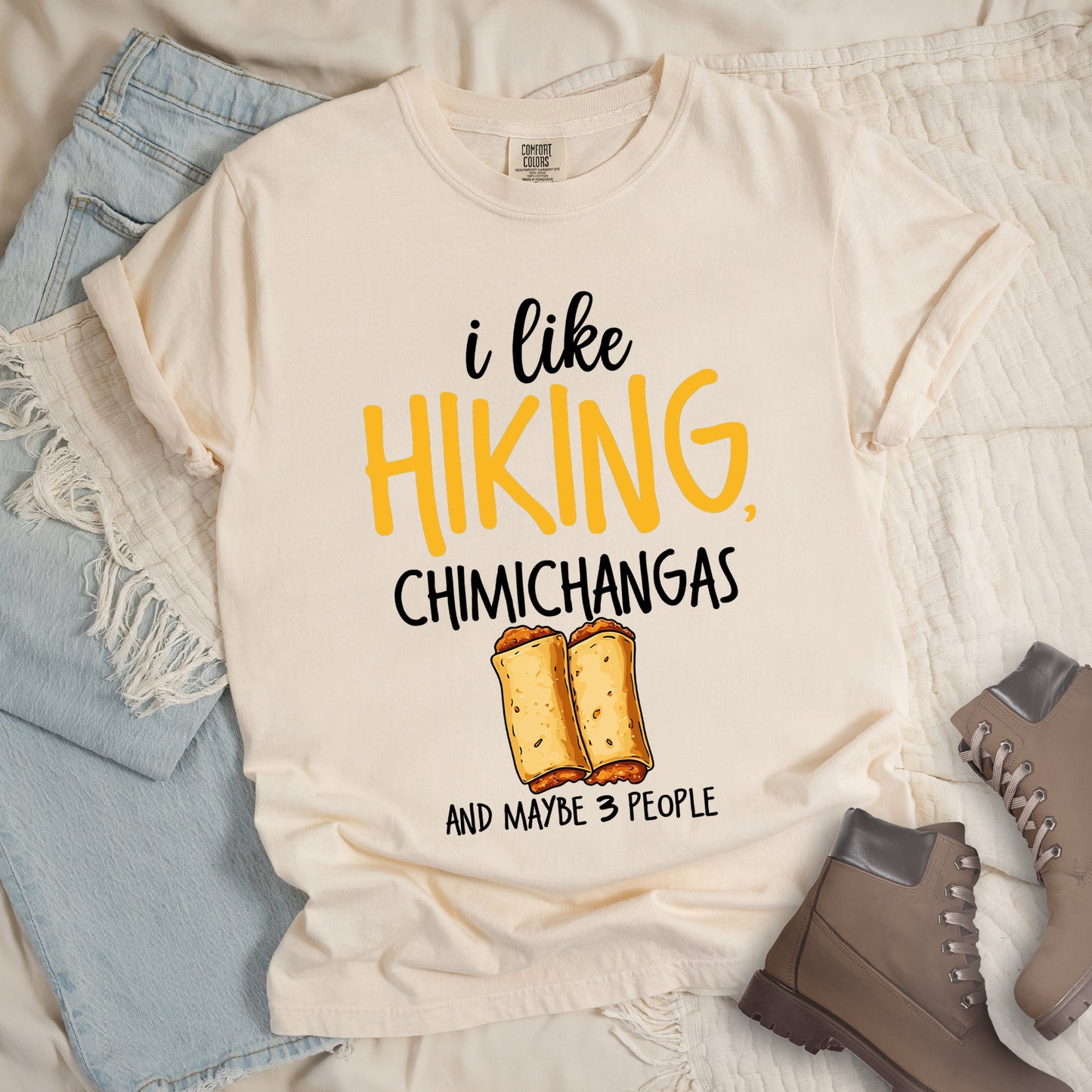Ivory (Cream White) t-shirt with text "i like HIKING, CHIMICHANGAS AND MAYBE 3 PEOPLE". The phrase "i like" appears in black script at the top, "HIKING," in yellow letters, "CHIMICHANGAS" in black, and "AND MAYBE 3 PEOPLE" in black at the bottom. Between "CHIMICHANGAS" and "AND MAYBE 3 PEOPLE" is an illustration of two chimichangas (fried burritos) with visible fillings. The design combines hiking, food, and introvert humor.