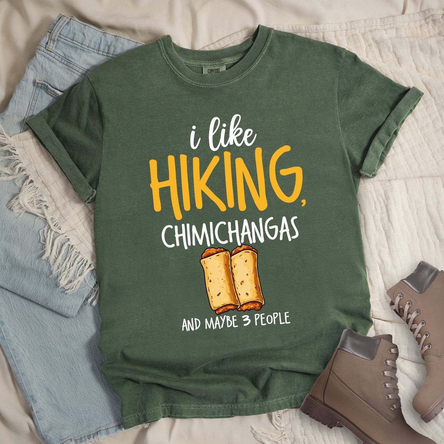 Moss (Dark Green) t-shirt with text "i like HIKING, CHIMICHANGAS AND MAYBE 3 PEOPLE". The phrase "i like" appears in white script at the top, "HIKING," in yellow letters, "CHIMICHANGAS" in white, and "AND MAYBE 3 PEOPLE" in white at the bottom. Between "CHIMICHANGAS" and "AND MAYBE 3 PEOPLE" is an illustration of two chimichangas (fried burritos) with visible fillings. The design combines hiking, food, and introvert humor.