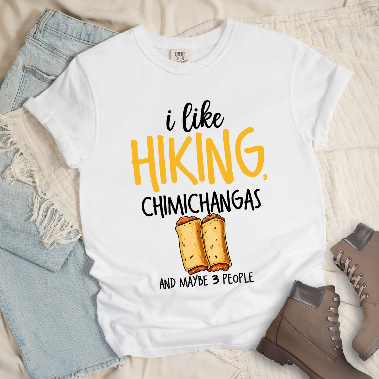 White t-shirt with text "i like HIKING, CHIMICHANGAS AND MAYBE 3 PEOPLE". The phrase "i like" appears in black script at the top, "HIKING," in yellow letters, "CHIMICHANGAS" in black, and "AND MAYBE 3 PEOPLE" in black at the bottom. Between "CHIMICHANGAS" and "AND MAYBE 3 PEOPLE" is an illustration of two chimichangas (fried burritos) with visible fillings. The design combines hiking, food, and introvert humor.