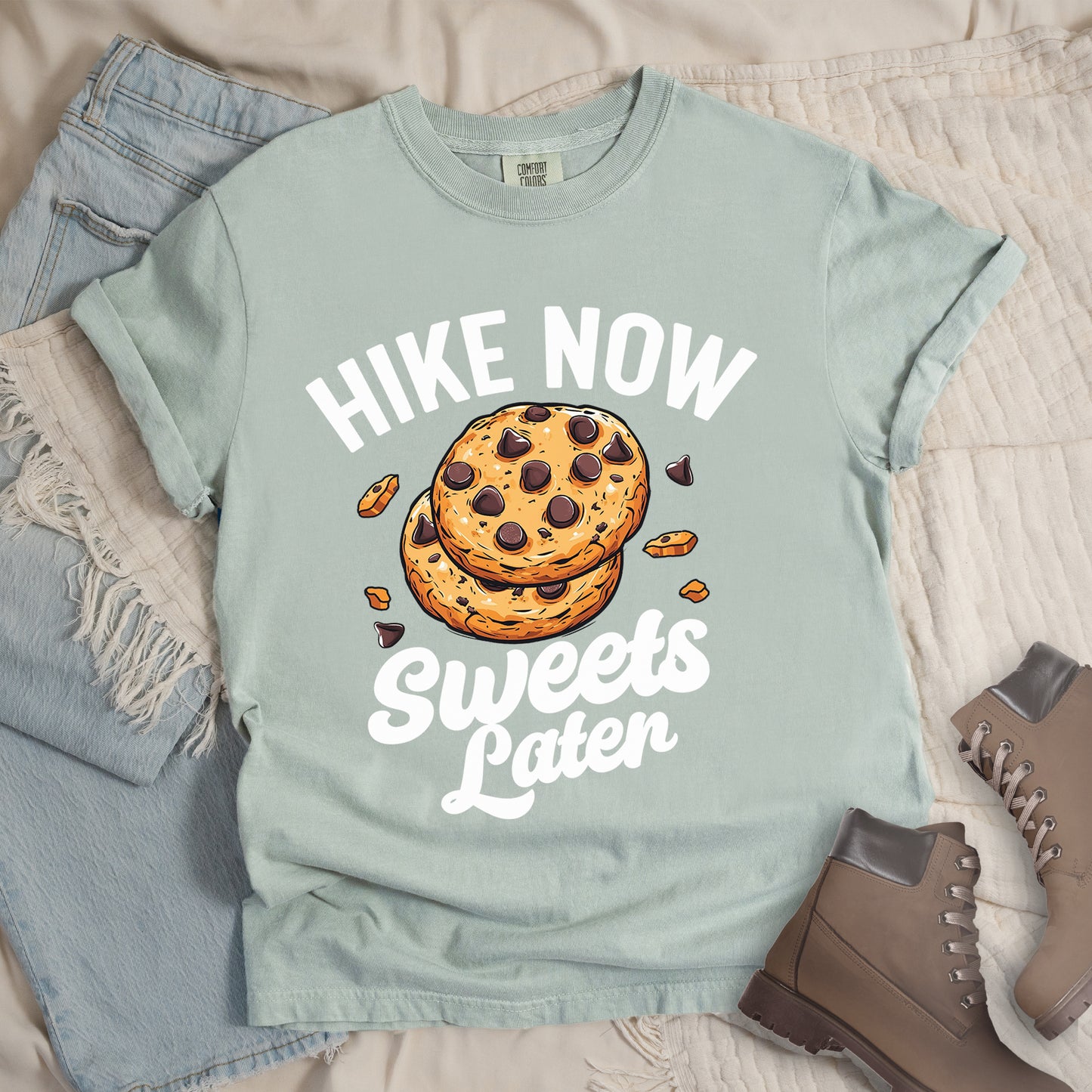 Bay (Light Blue) t-shirt with "HIKE NOW Sweets Later" text design. "HIKE NOW" appears in white capital letters arched at the top, and "Sweets Later" in white script at the bottom. In the center is an illustration of two chocolate chip cookies stacked on top of each other with some cookie crumbs and chocolate chunks scattered around them. The design emphasizes hiking first with dessert as a reward afterward.