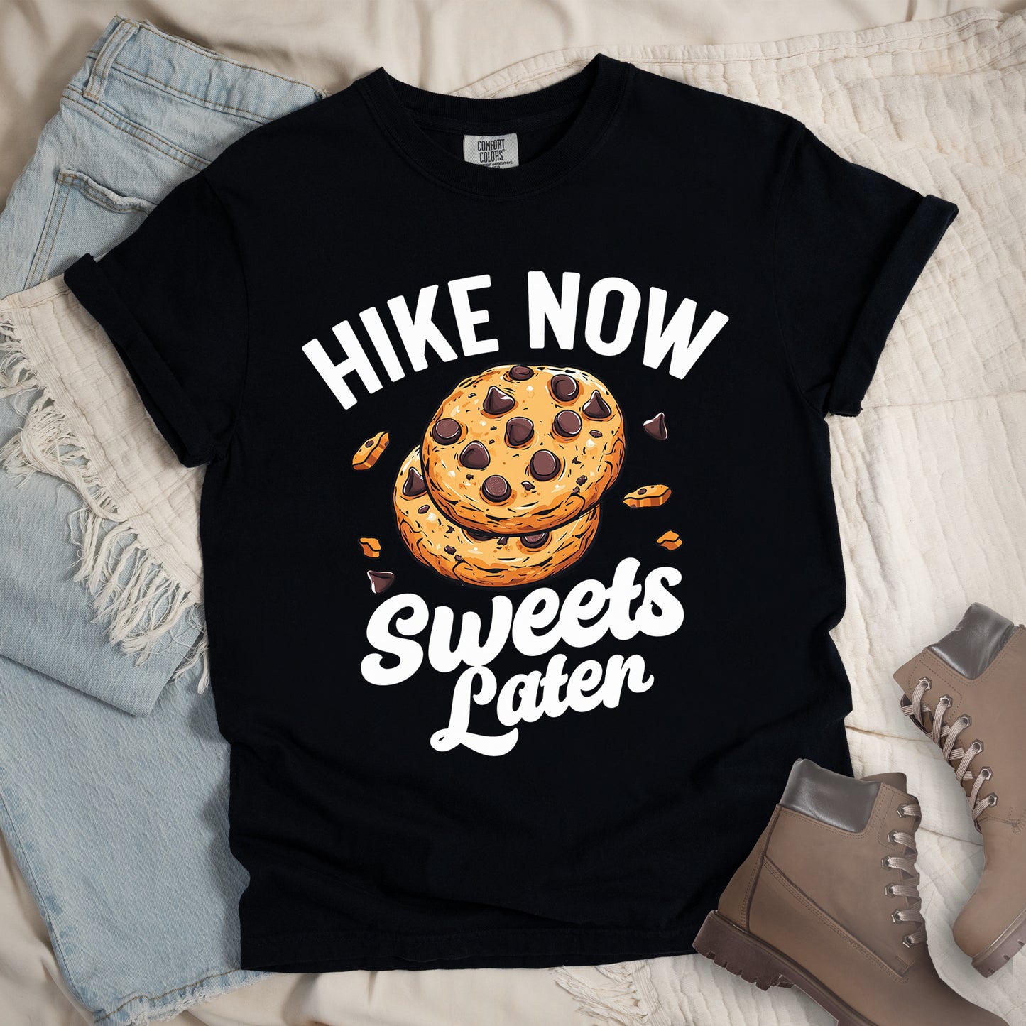 Black t-shirt with "HIKE NOW Sweets Later" text design. "HIKE NOW" appears in white capital letters arched at the top, and "Sweets Later" in white script at the bottom. In the center is an illustration of two chocolate chip cookies stacked on top of each other with some cookie crumbs and chocolate chunks scattered around them. The design emphasizes hiking first with dessert as a reward afterward.