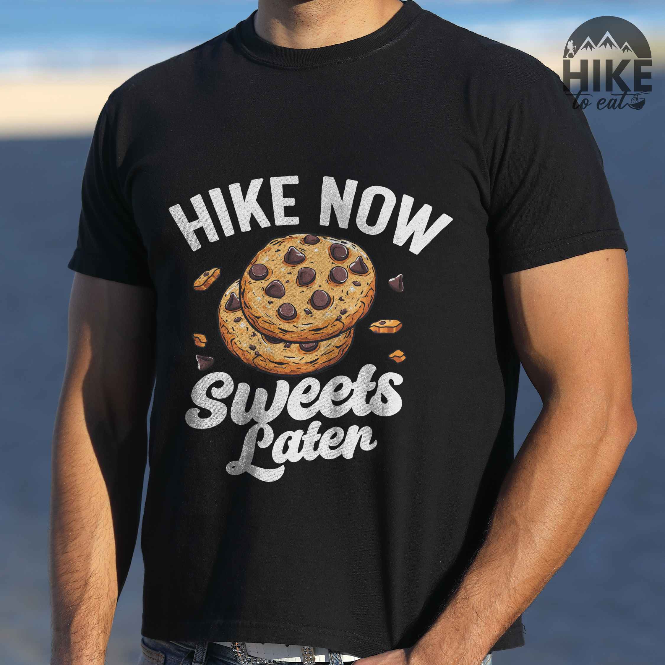 Man wearing black “Hike Now Sweets Later” colored vintage tee with bold and script typography, illustration of chocolate chip cookies with crumbs scattered around. Perfect for cookie lovers.