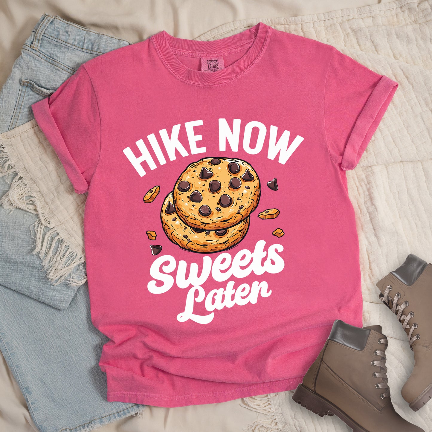 Crunchberry (Bright Pink) t-shirt with "HIKE NOW Sweets Later" text design. "HIKE NOW" appears in white capital letters arched at the top, and "Sweets Later" in white script at the bottom. In the center is an illustration of two chocolate chip cookies stacked on top of each other with some cookie crumbs and chocolate chunks scattered around them. The design emphasizes hiking first with dessert as a reward afterward.
