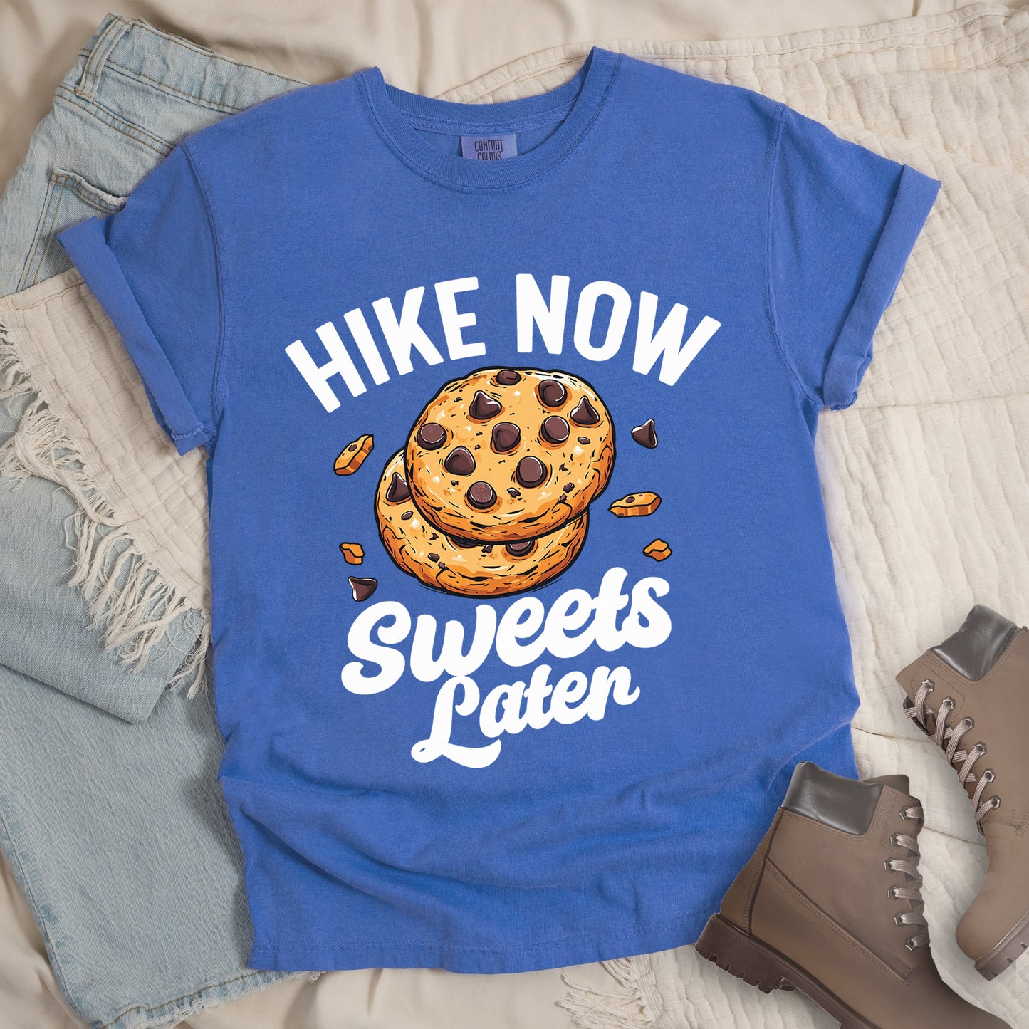 Flo Blue (Bright Blue) t-shirt with "HIKE NOW Sweets Later" text design. "HIKE NOW" appears in white capital letters arched at the top, and "Sweets Later" in white script at the bottom. In the center is an illustration of two chocolate chip cookies stacked on top of each other with some cookie crumbs and chocolate chunks scattered around them. The design emphasizes hiking first with dessert as a reward afterward.