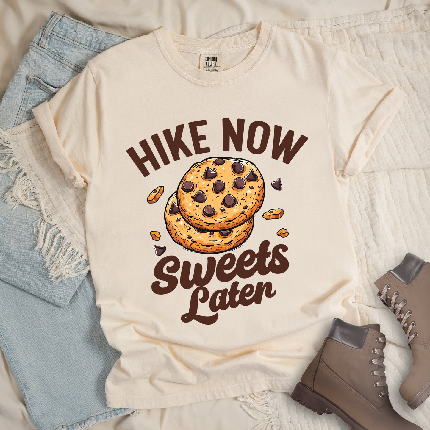 Ivory (Cream White) t-shirt with "HIKE NOW Sweets Later" text design. "HIKE NOW" appears in brown capital letters arched at the top, and "Sweets Later" in brown script at the bottom. In the center is an illustration of two chocolate chip cookies stacked on top of each other with some cookie crumbs and chocolate chunks scattered around them. The design emphasizes hiking first with dessert as a reward afterward.