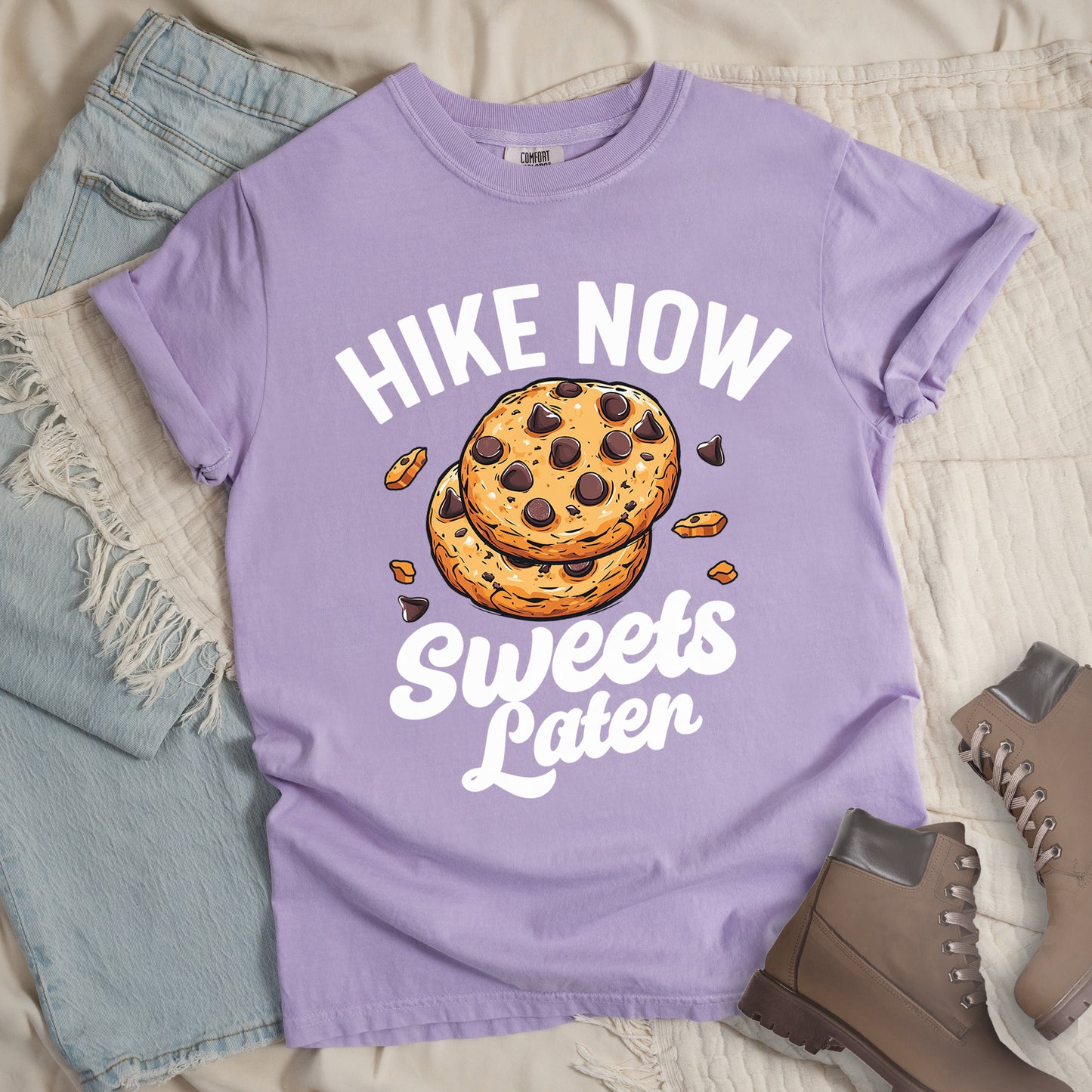 Orchid (Light Violet) t-shirt with "HIKE NOW Sweets Later" text design. "HIKE NOW" appears in white capital letters arched at the top, and "Sweets Later" in white script at the bottom. In the center is an illustration of two chocolate chip cookies stacked on top of each other with some cookie crumbs and chocolate chunks scattered around them. The design emphasizes hiking first with dessert as a reward afterward.