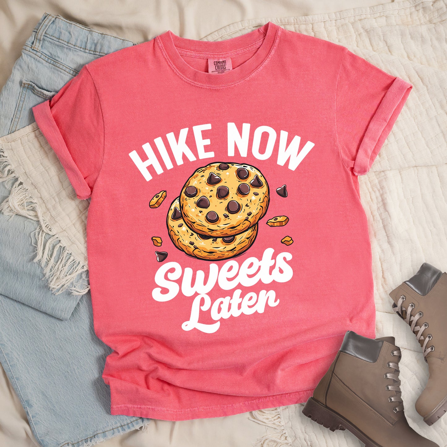 Watermelon (Light Coral) t-shirt with "HIKE NOW Sweets Later" text design. "HIKE NOW" appears in white capital letters arched at the top, and "Sweets Later" in white script at the bottom. In the center is an illustration of two chocolate chip cookies stacked on top of each other with some cookie crumbs and chocolate chunks scattered around them. The design emphasizes hiking first with dessert as a reward afterward.