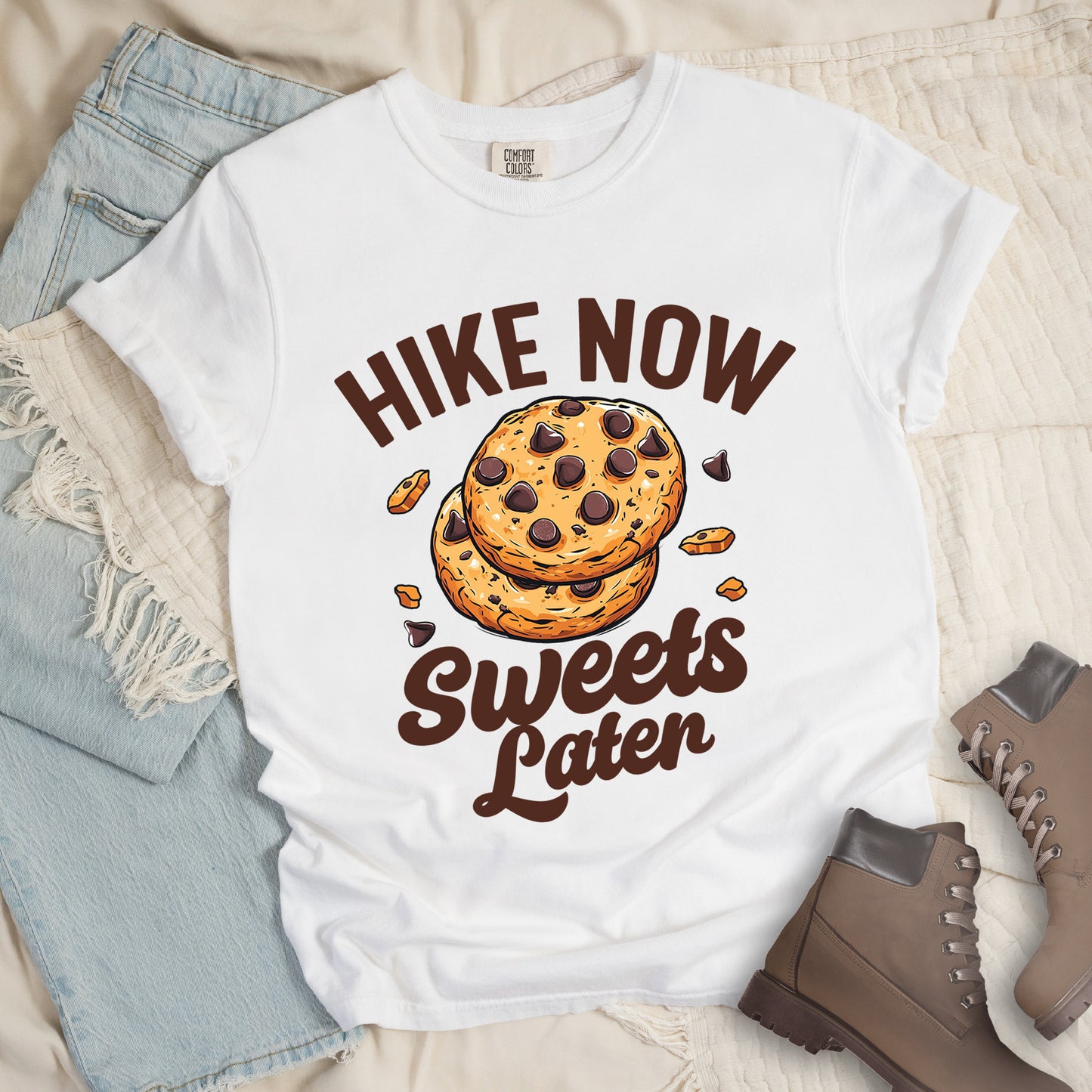 White t-shirt with "HIKE NOW Sweets Later" text design. "HIKE NOW" appears in brown capital letters arched at the top, and "Sweets Later" in brown script at the bottom. In the center is an illustration of two chocolate chip cookies stacked on top of each other with some cookie crumbs and chocolate chunks scattered around them. The design emphasizes hiking first with dessert as a reward afterward.