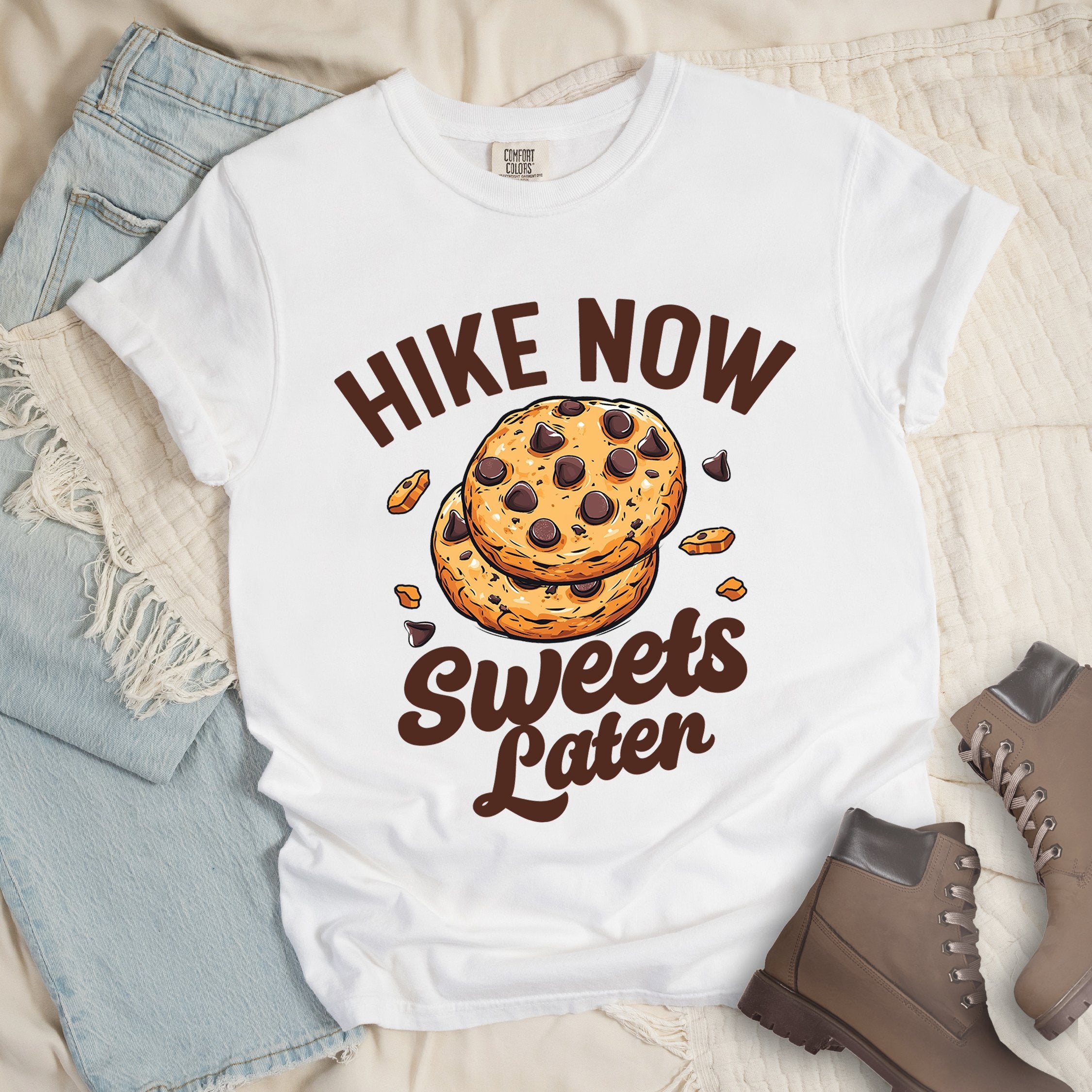 White shirt with Hiking Design featuring the phrase “Hike Now Sweets Later” and an illustration of chocolate chip cookies with crumbs scattered around.