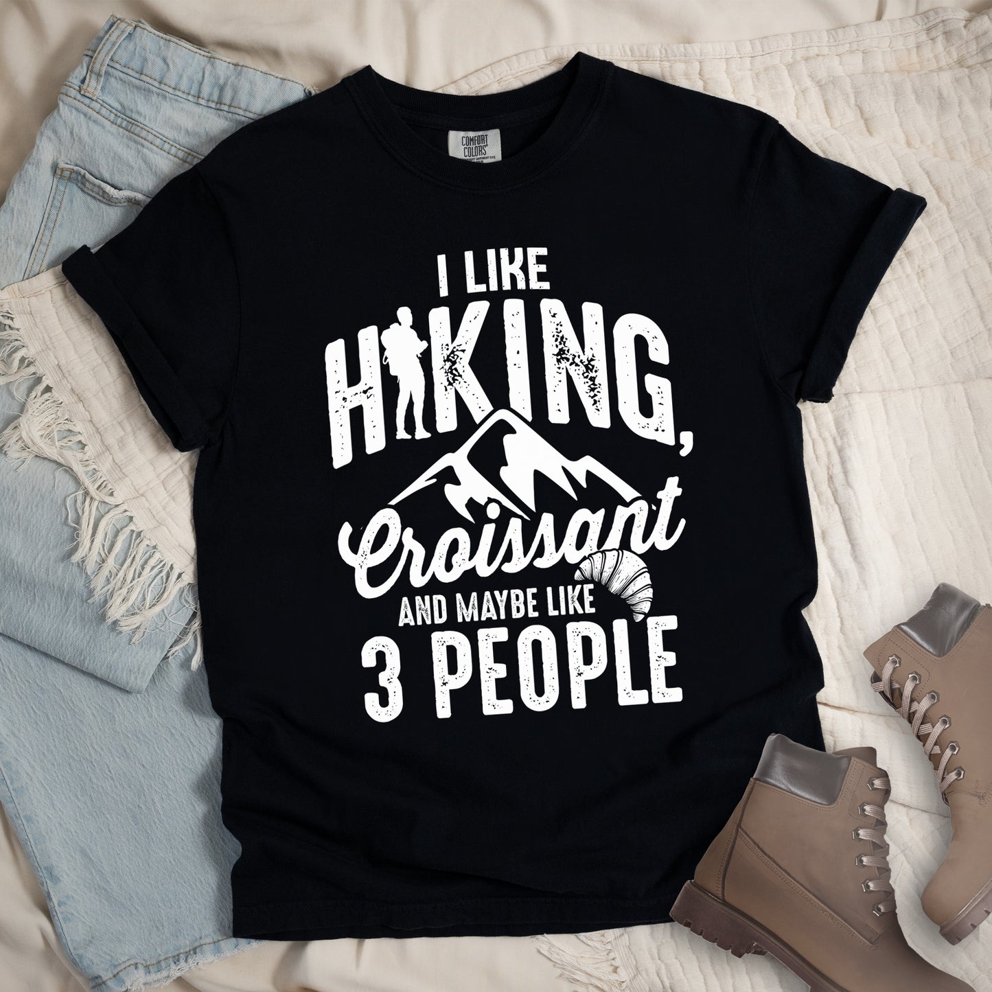Black t-shirt with distressed white text reading "I LIKE HIKING, Croissant AND MAYBE LIKE 3 PEOPLE". The letter "I" in "HIKING" is replaced with a hiker silhouette. Below "HIKING," are mountain silhouettes, followed by "Croissant" in script with a small croissant illustration. The design combines outdoor activity, food preference, and introvert humor in a minimalist white-on-black style. Features a classic crew neck and casual rolled sleeves, styled alongside light wash denim and brown hiking boots.