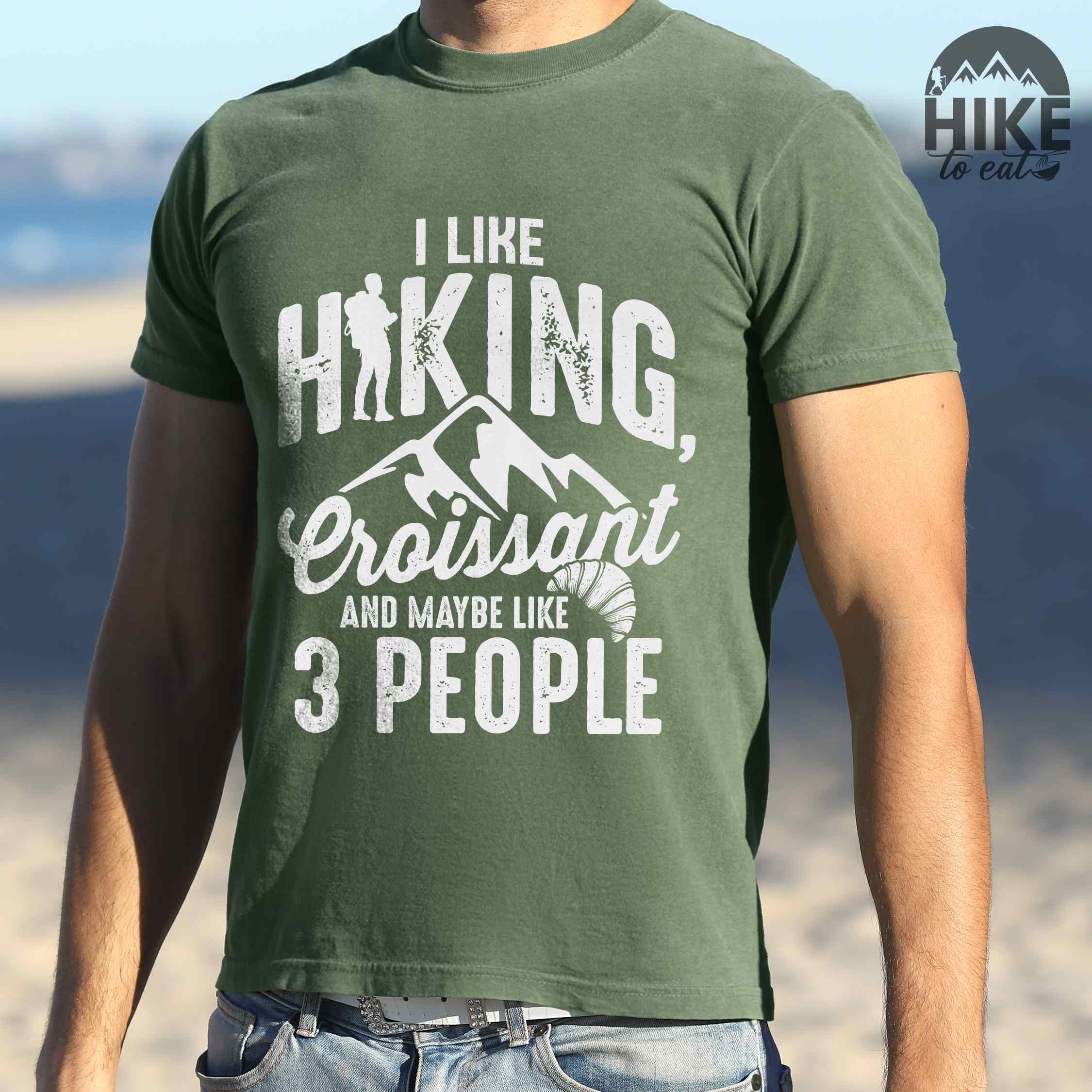 Man wearing moss "I Like hiking, croissant and maybe like 3 people" colored vintage tee with bold typography, birds, mountains, illustrated croissant, and backpacker for pastry enthusiasts.