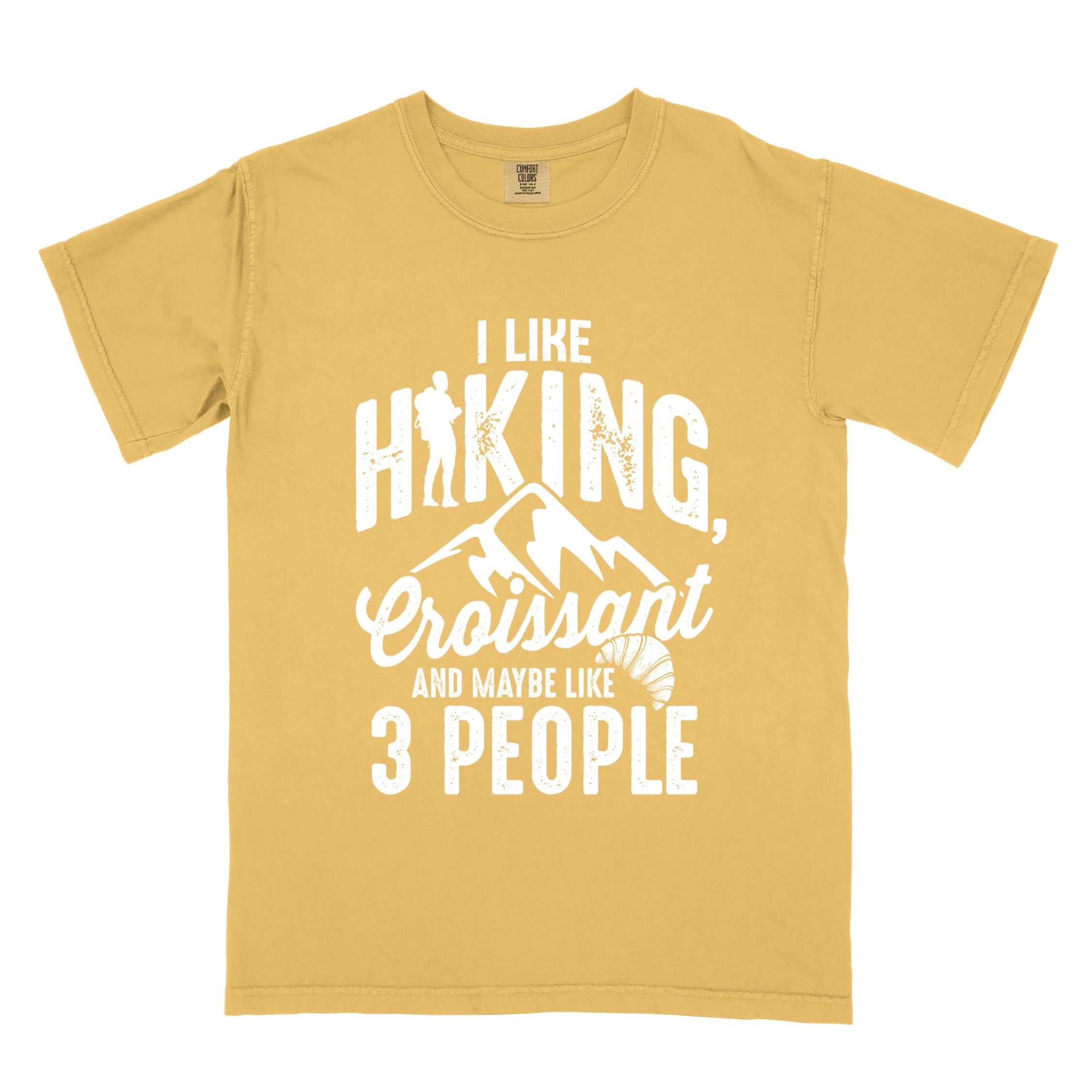 Mustard "I Like hiking, croissant and maybe like 3 people" colored vintage tee with bold typography, birds, mountains, illustrated croissant, and backpacker for pastry enthusiasts.