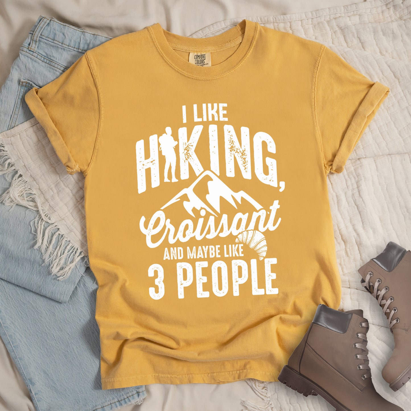 Mustard (Yellow) t-shirt with distressed white text reading "I LIKE HIKING, Croissant AND MAYBE LIKE 3 PEOPLE". The letter "I" in "HIKING" is replaced with a hiker silhouette. Below "HIKING," are mountain silhouettes, followed by "Croissant" in script with a small croissant illustration. The design combines outdoor activity, food preference, and introvert humor in a minimalist white-on-yellow style.