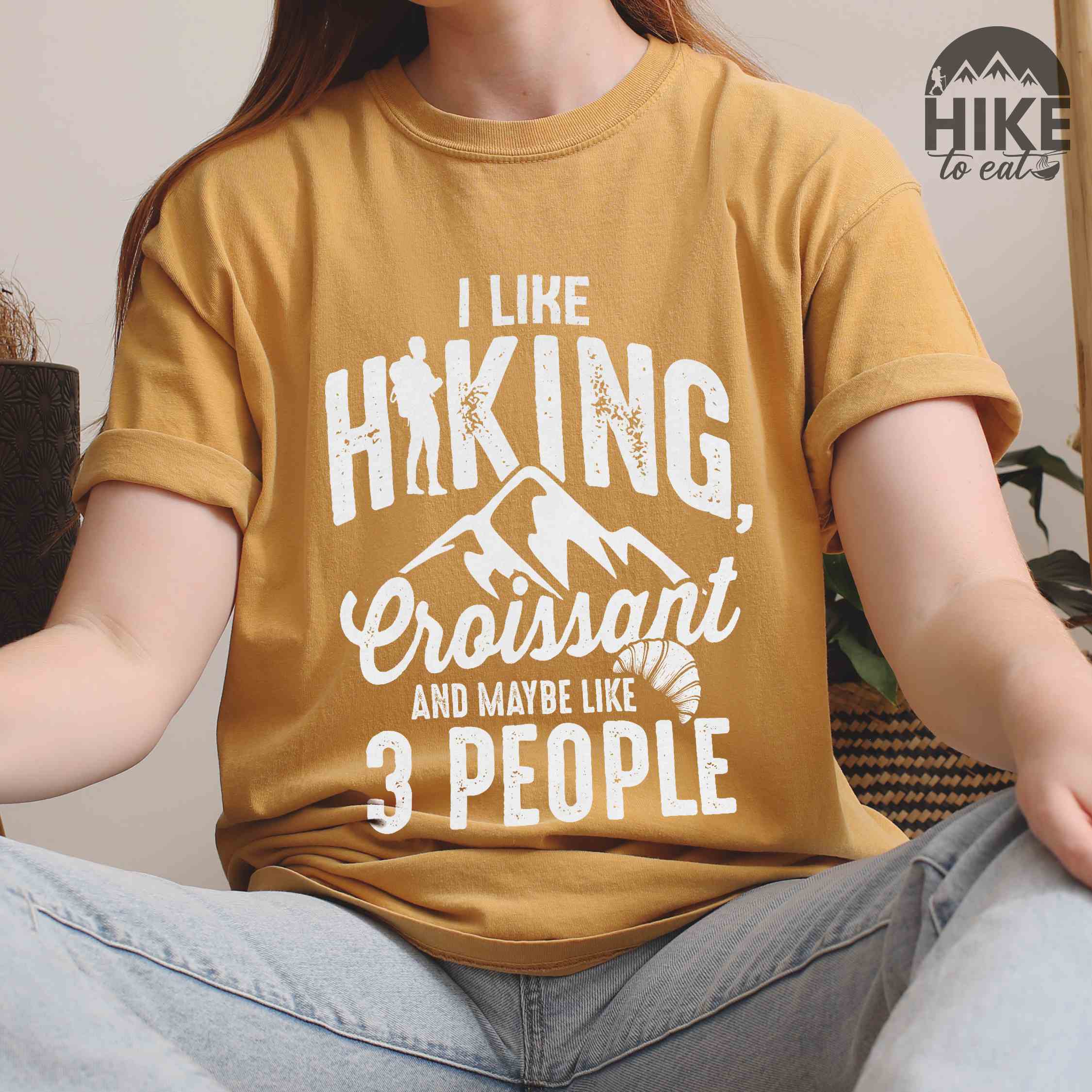 woman wearing mustard "I Like hiking, croissant and maybe like 3 people" colored vintage tee with bold typography, birds, mountains, illustrated croissant, and backpacker for pastry enthusiasts.