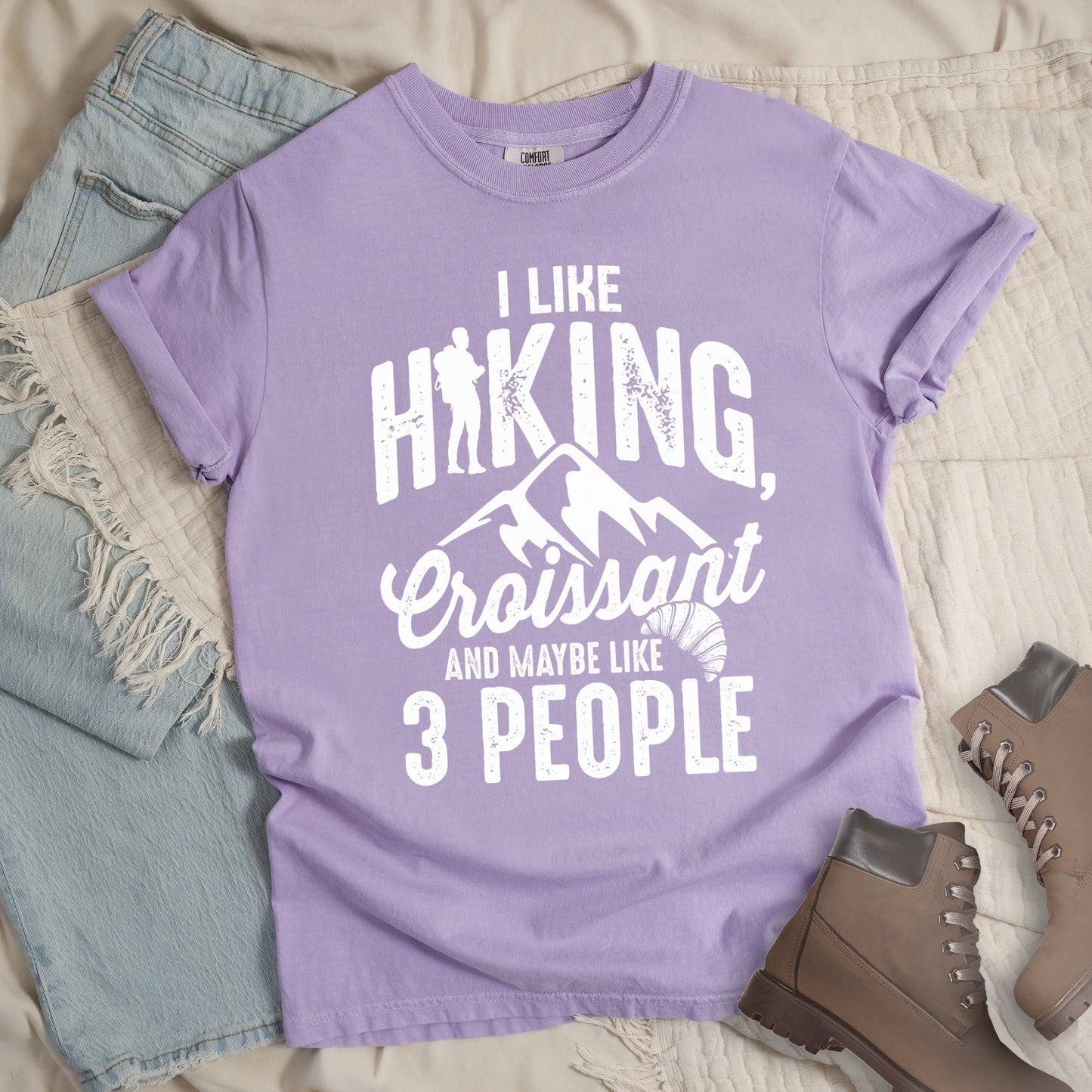 Orchid (Light Violet) t-shirt with distressed white text reading "I LIKE HIKING, Croissant AND MAYBE LIKE 3 PEOPLE". The letter "I" in "HIKING" is replaced with a hiker silhouette. Below "HIKING," are mountain silhouettes, followed by "Croissant" in script with a small croissant illustration. The design combines outdoor activity, food preference, and introvert humor in a minimalist white-on-purple style.