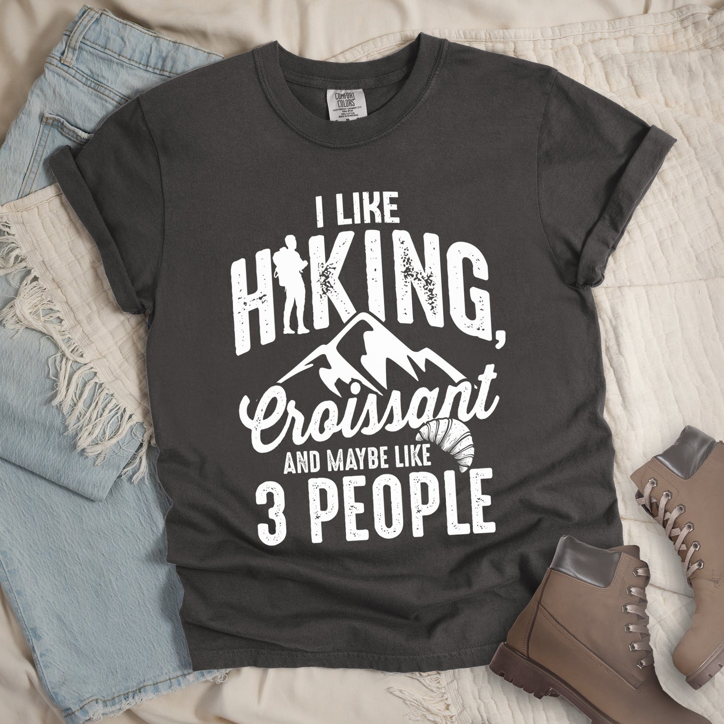 Pepper (Dark Gray) t-shirt with distressed white text reading "I LIKE HIKING, Croissant AND MAYBE LIKE 3 PEOPLE". The letter "I" in "HIKING" is replaced with a hiker silhouette. Below "HIKING," are mountain silhouettes, followed by "Croissant" in script with a small croissant illustration. The design combines outdoor activity, food preference, and introvert humor in a minimalist white-on-gray style.