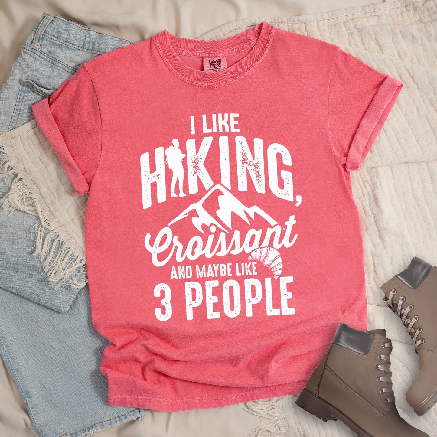 Watermelon (Light Coral) t-shirt with distressed white text reading "I LIKE HIKING, Croissant AND MAYBE LIKE 3 PEOPLE". The letter "I" in "HIKING" is replaced with a hiker silhouette. Below "HIKING," are mountain silhouettes, followed by "Croissant" in script with a small croissant illustration. The design combines outdoor activity, food preference, and introvert humor in a minimalist white-on-pink style.