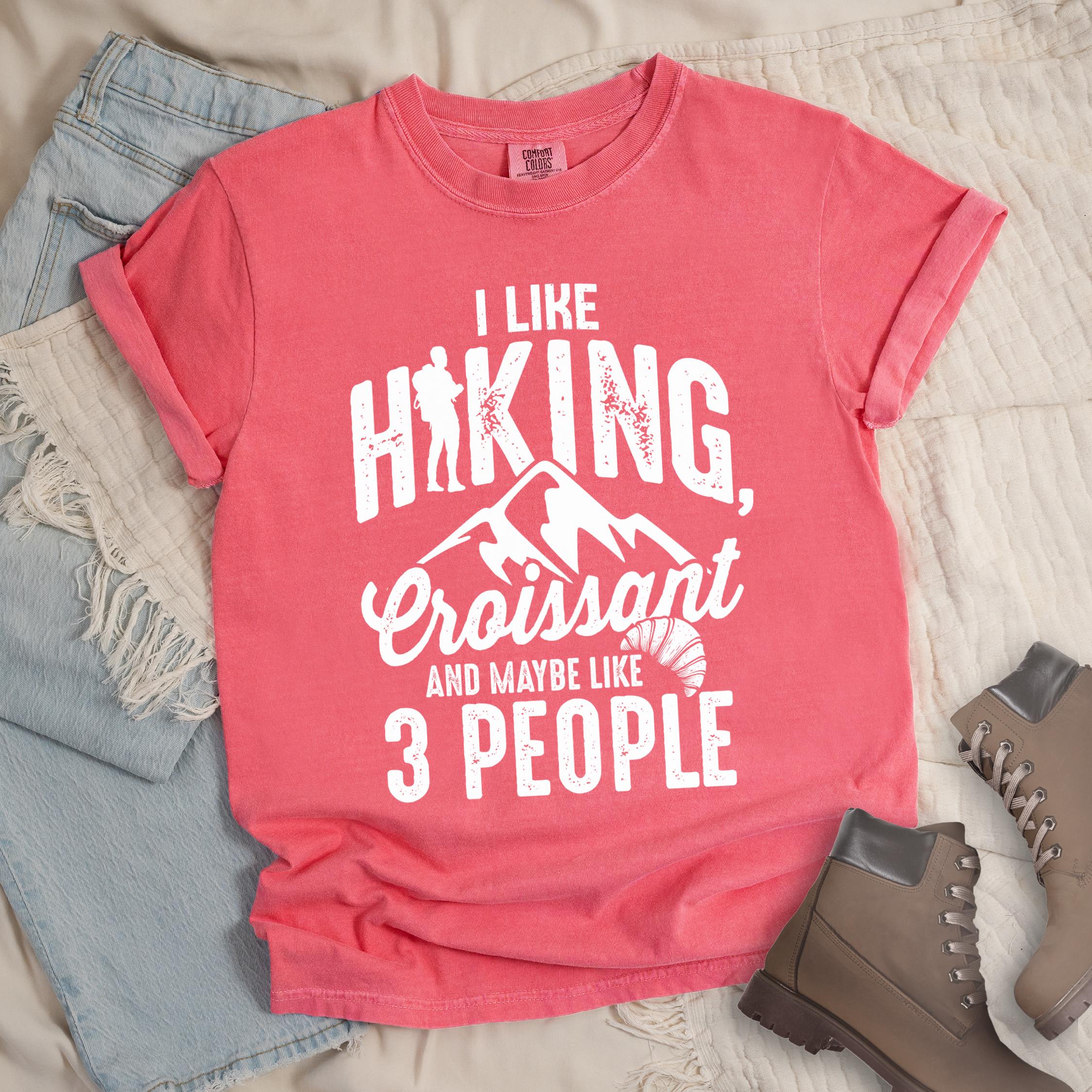 Pink shirt with Hiking Design featuring the phrase “I Like Hiking, Croissant, and Maybe Like 3 People” and an illustration of a croissant below mountain outlines.