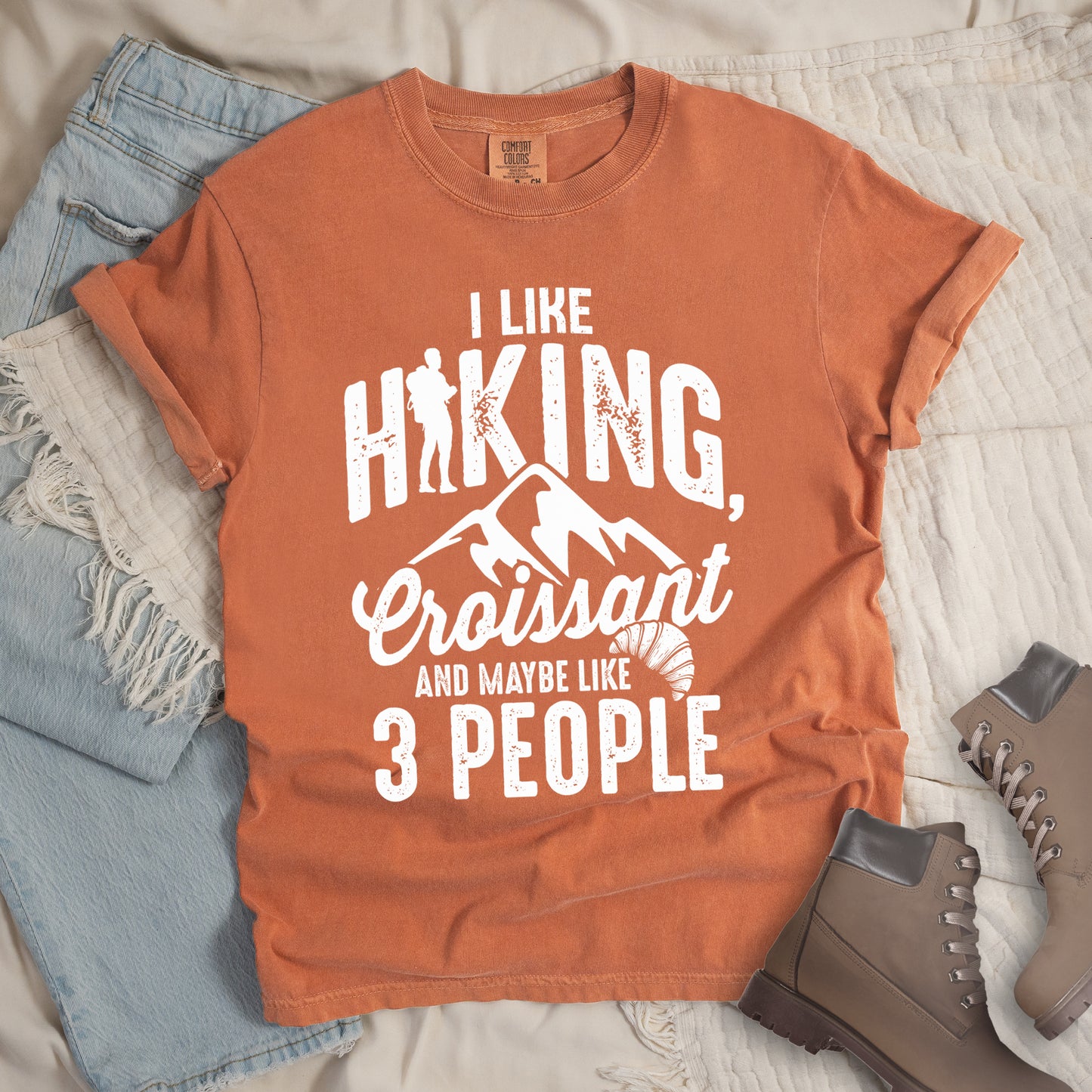 Yam (Brown) t-shirt with distressed white text reading "I LIKE HIKING, Croissant AND MAYBE LIKE 3 PEOPLE". The letter "I" in "HIKING" is replaced with a hiker silhouette. Below "HIKING," are mountain silhouettes, followed by "Croissant" in script with a small croissant illustration. The design combines outdoor activity, food preference, and introvert humor in a minimalist white-on-brown style.