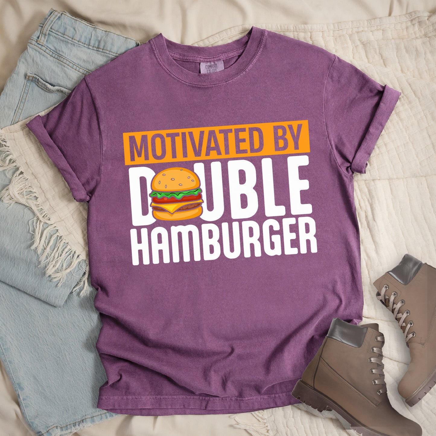 Berry (Dark Violet) t-shirt with text "MOTIVATED BY DOUBLE HAMBURGER". "MOTIVATED BY" appears in Berry (Dark Violet) text on an orange rectangular background at the top. "DOUBLE HAMBURGER" appears in large white letters below, with a colorful hamburger illustration replacing the letter "O" in "DOUBLE". The hamburger graphic shows a sesame seed bun, lettuce, tomato, cheese, and beef patty.