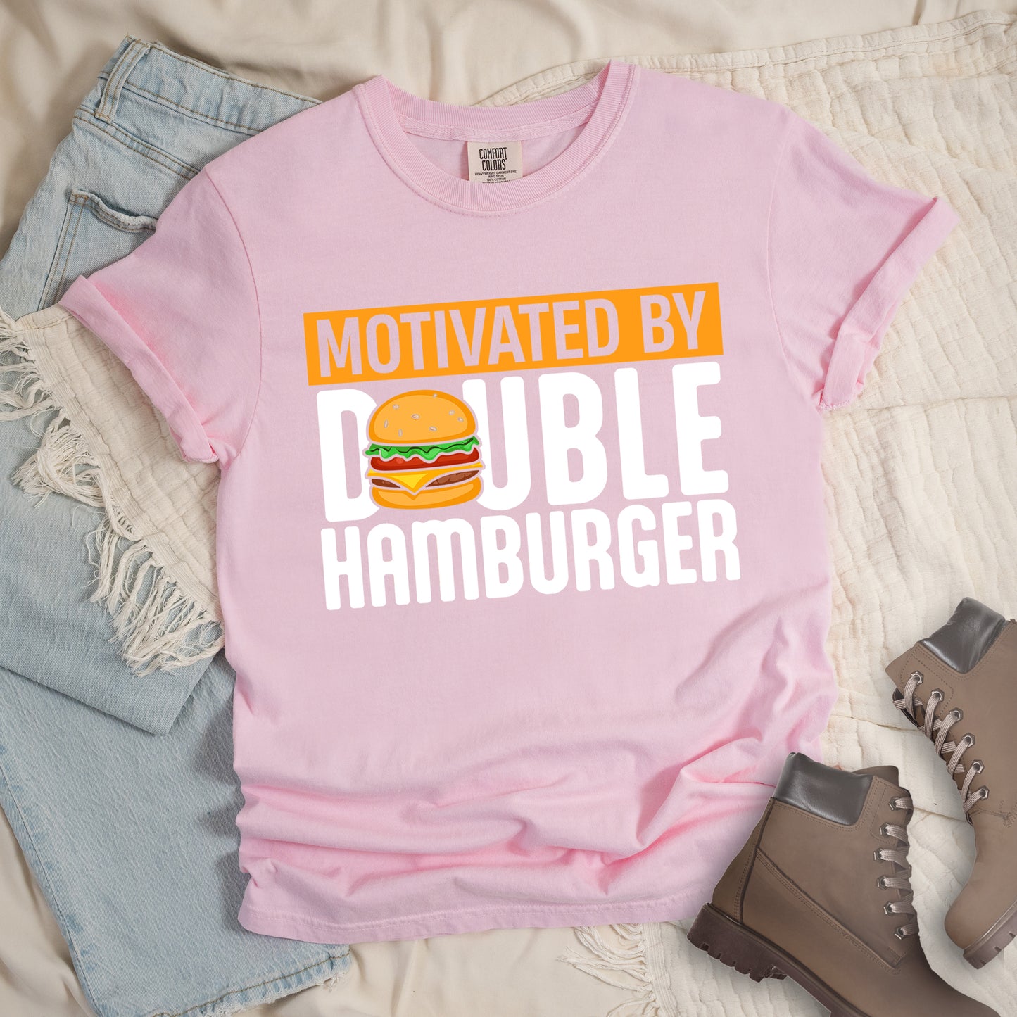 Blossom (Light Pink) t-shirt with text "MOTIVATED BY DOUBLE HAMBURGER". "MOTIVATED BY" appears in Blossom (Light Pink) text on an orange rectangular background at the top. "DOUBLE HAMBURGER" appears in large white letters below, with a colorful hamburger illustration replacing the letter "O" in "DOUBLE". The hamburger graphic shows a sesame seed bun, lettuce, tomato, cheese, and beef patty.