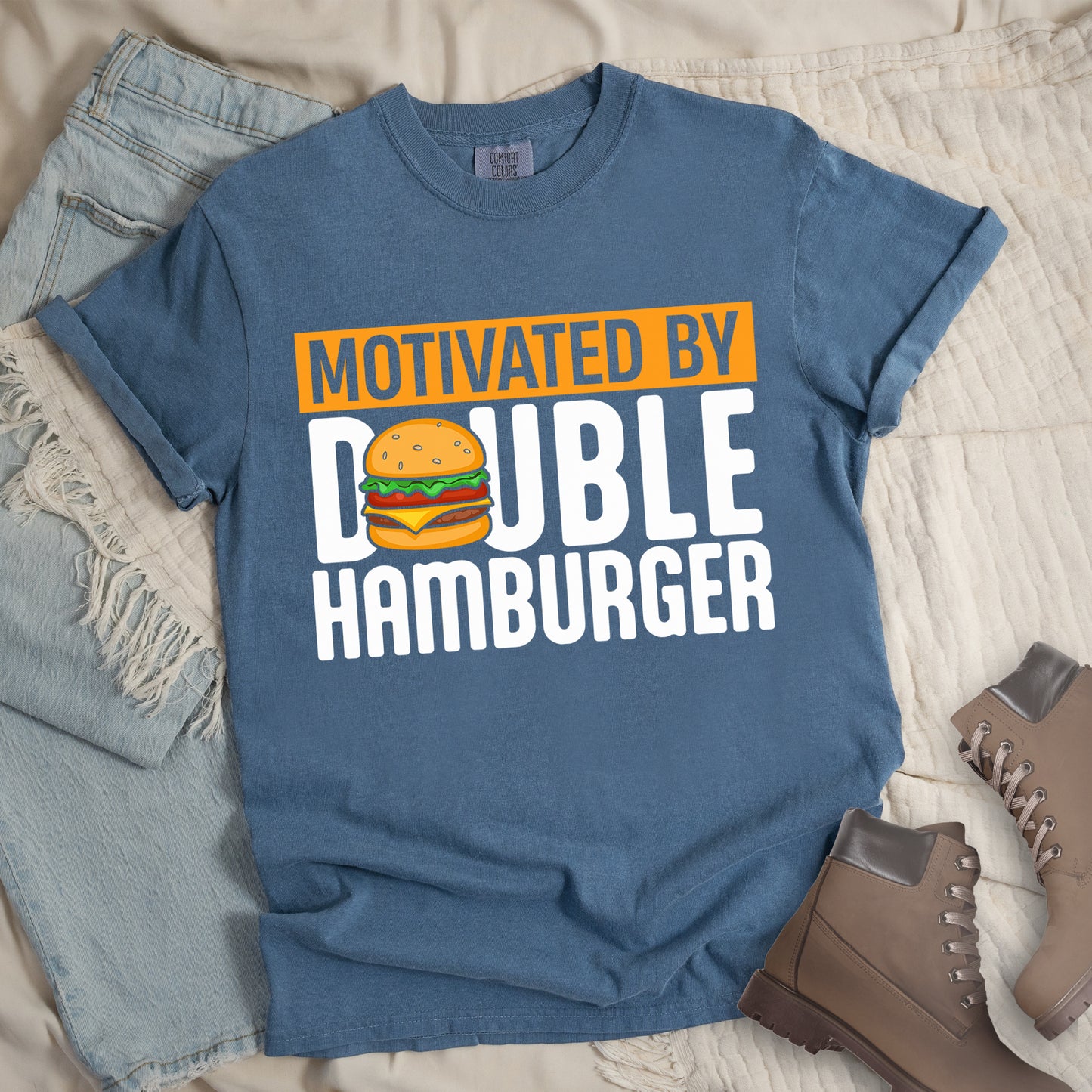 Blue Jean (Denim Blue) t-shirt with text "MOTIVATED BY DOUBLE HAMBURGER". "MOTIVATED BY" appears in Blue Jean (Denim Blue) text on an orange rectangular background at the top. "DOUBLE HAMBURGER" appears in large white letters below, with a colorful hamburger illustration replacing the letter "O" in "DOUBLE". The hamburger graphic shows a sesame seed bun, lettuce, tomato, cheese, and beef patty.