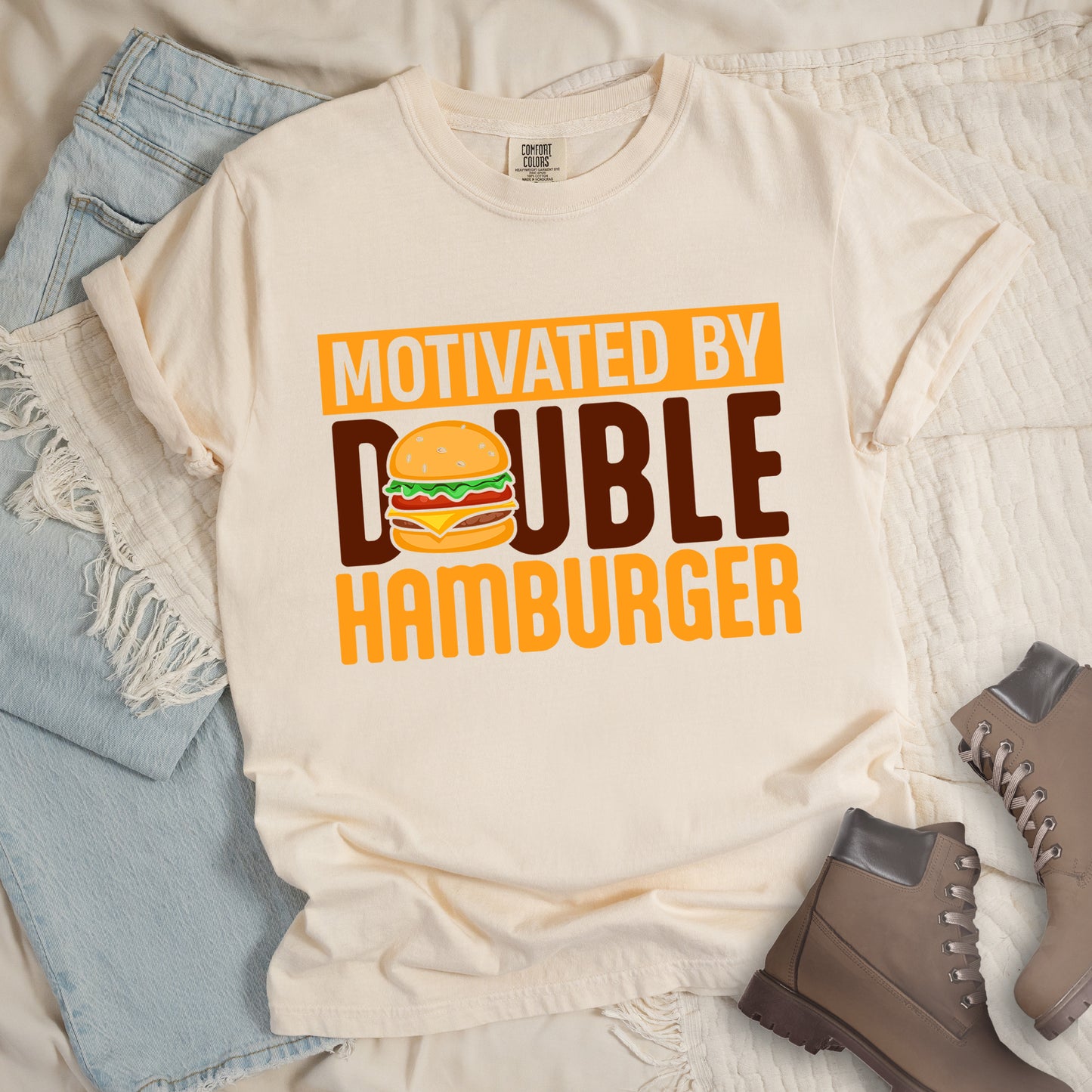 Ivory (Cream White) t-shirt with text "MOTIVATED BY DOUBLE HAMBURGER". "MOTIVATED BY" appears in Ivory (Cream White) text on an orange rectangular background at the top. "DOUBLE HAMBURGER" appears in large deep brown and yellow letters below, with a colorful hamburger illustration replacing the letter "O" in "DOUBLE". The hamburger graphic shows a sesame seed bun, lettuce, tomato, cheese, and beef patty.