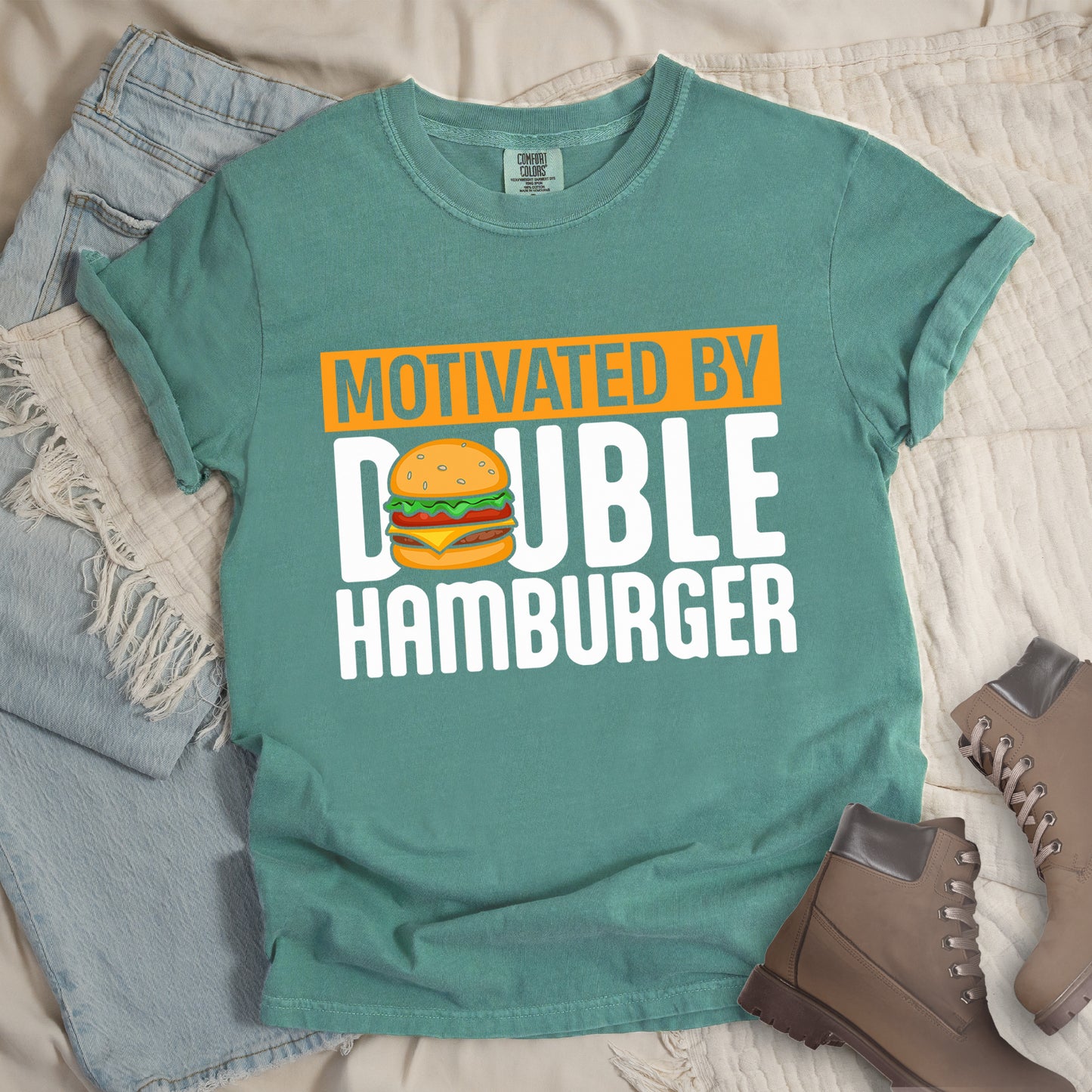 Light Green t-shirt with text "MOTIVATED BY DOUBLE HAMBURGER". "MOTIVATED BY" appears in Light Green text on an orange rectangular background at the top. "DOUBLE HAMBURGER" appears in large white letters below, with a colorful hamburger illustration replacing the letter "O" in "DOUBLE". The hamburger graphic shows a sesame seed bun, lettuce, tomato, cheese, and beef patty.