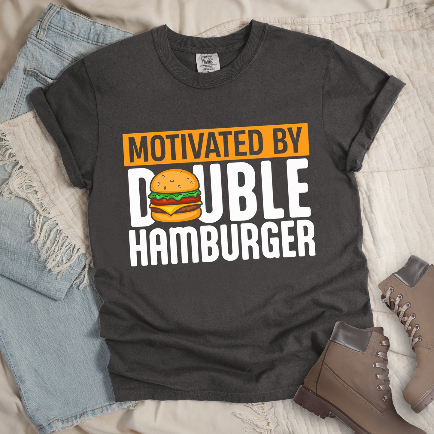 Pepper (Dark Gray) t-shirt with text "MOTIVATED BY DOUBLE HAMBURGER". "MOTIVATED BY" appears in Pepper (Dark Gray) text on an orange rectangular background at the top. "DOUBLE HAMBURGER" appears in large white letters below, with a colorful hamburger illustration replacing the letter "O" in "DOUBLE". The hamburger graphic shows a sesame seed bun, lettuce, tomato, cheese, and beef patty.