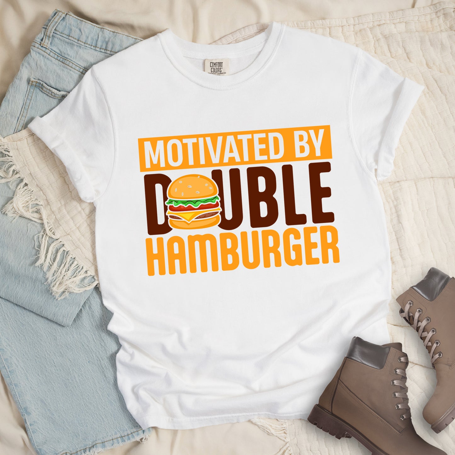 White t-shirt with text "MOTIVATED BY DOUBLE HAMBURGER". "MOTIVATED BY" appears in white text on an orange rectangular background at the top. "DOUBLE HAMBURGER" appears in large deep brown and yellow letters below, with a colorful hamburger illustration replacing the letter "O" in "DOUBLE". The hamburger graphic shows a sesame seed bun, lettuce, tomato, cheese, and beef patty.