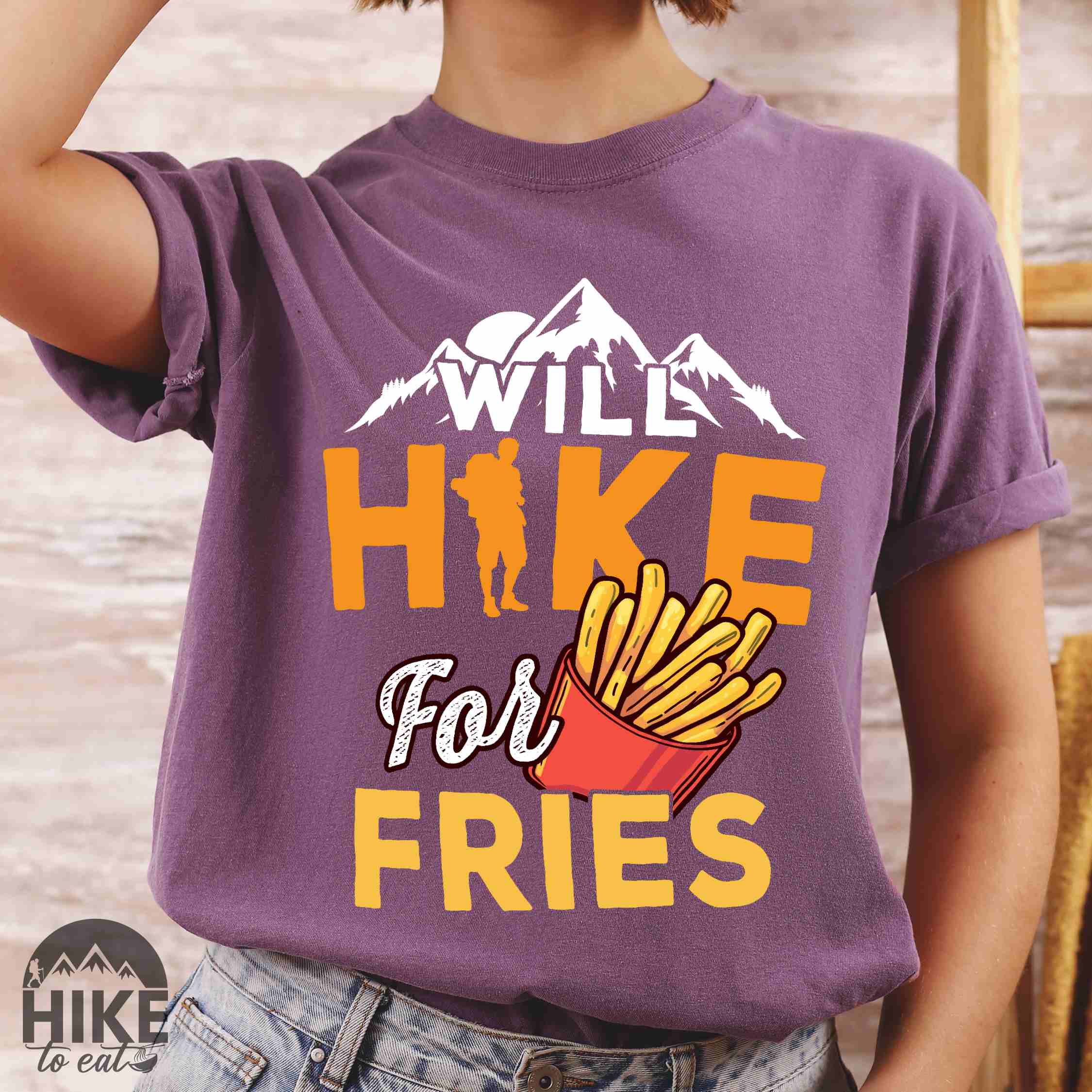 Woman wearing "Will Hike for Fries" colored vintage tee with bold typography, mountains, illustrated fries, and backpacker for comfort food enthusiasts.