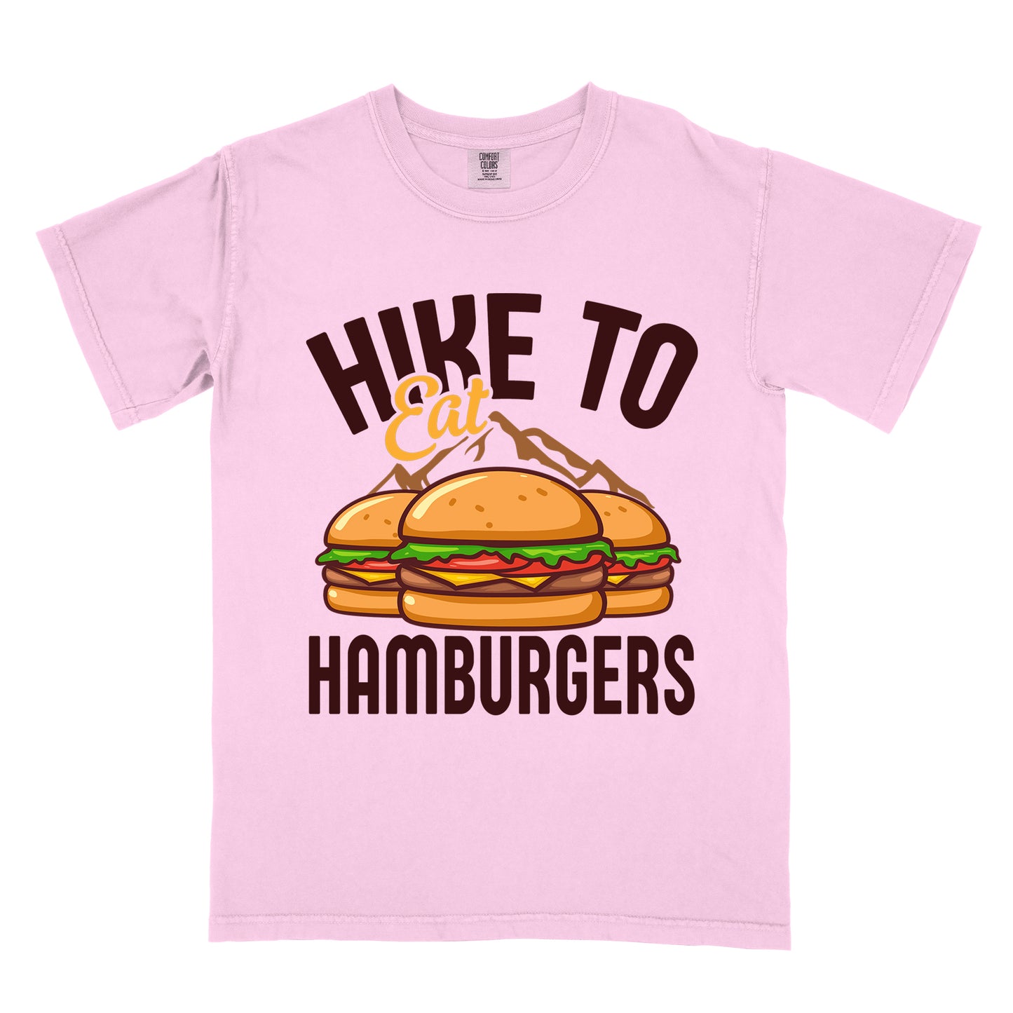 Blossom (Light Pink) t-shirt with text "HIKE TO Eat HAMBURGERS". "HIKE TO" appears in deep brown arched letters at the top, "Eat" in orange script in the middle, and "HAMBURGERS" in deep brown at the bottom. The center features an illustration of three hamburgers with buns, lettuce, tomato, cheese and beef patties, with brown mountain silhouettes behind them. The design connects hiking activity with food rewards.