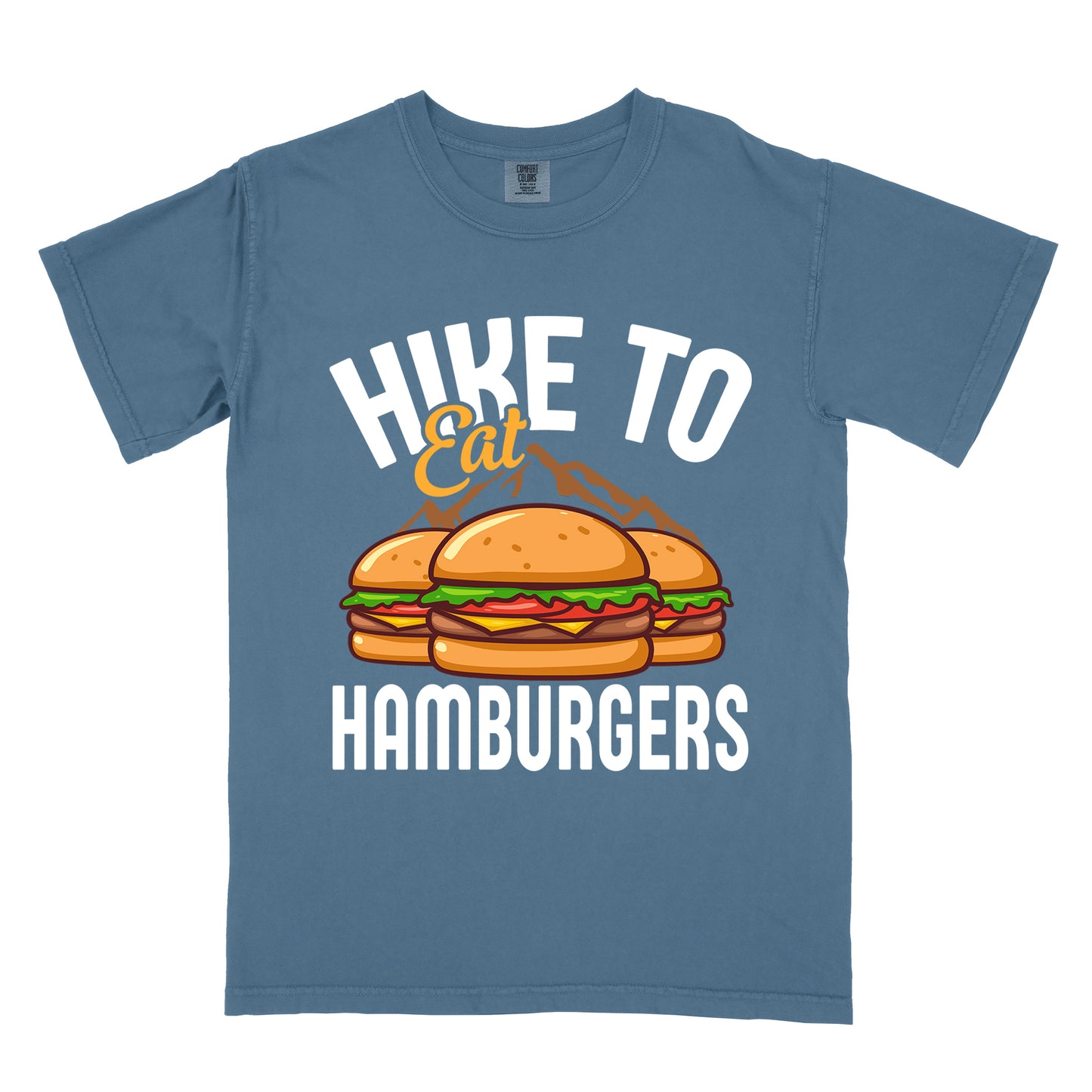 Blue Jean (Denim Blue) t-shirt with text "HIKE TO Eat HAMBURGERS". "HIKE TO" appears in white arched letters at the top, "Eat" in orange script in the middle, and "HAMBURGERS" in white at the bottom. The center features an illustration of three hamburgers with buns, lettuce, tomato, cheese and beef patties, with brown mountain silhouettes behind them. The design connects hiking activity with food rewards.
