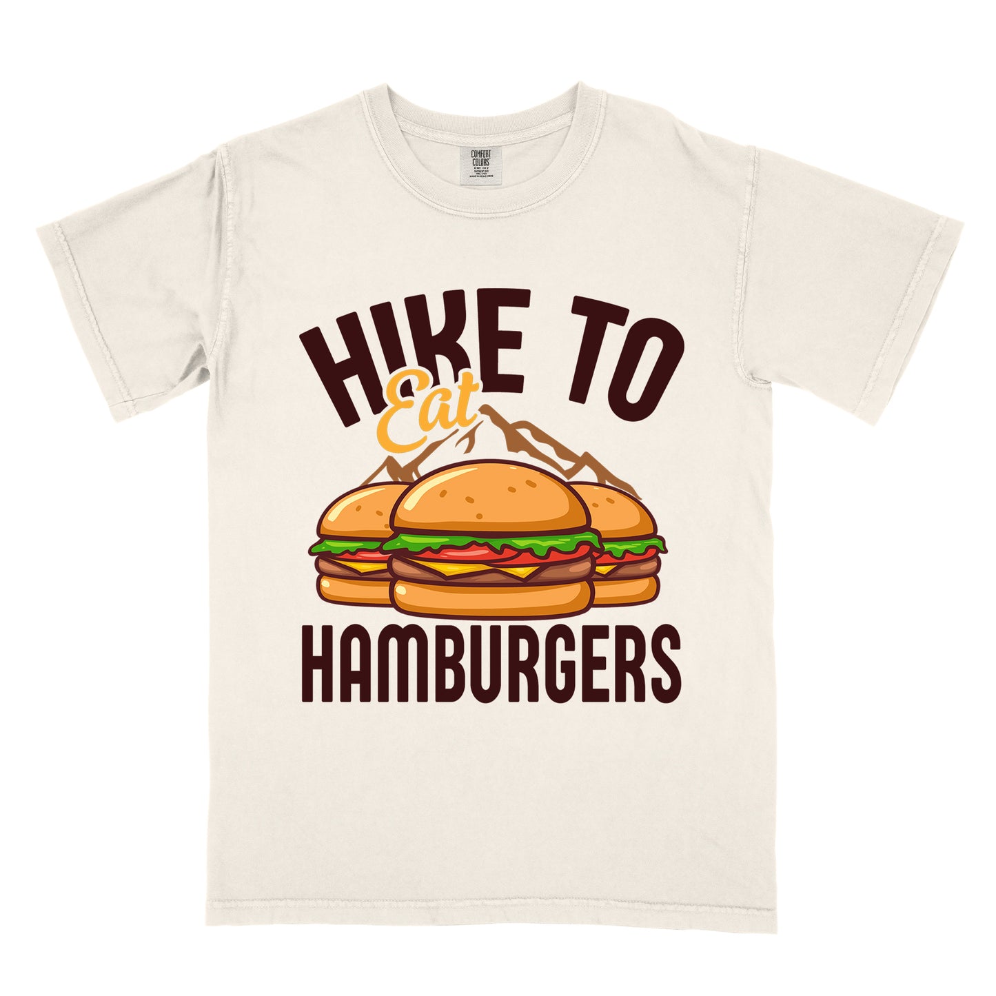 Ivory (Cream White) t-shirt with text "HIKE TO Eat HAMBURGERS". "HIKE TO" appears in deep brown arched letters at the top, "Eat" in orange script in the middle, and "HAMBURGERS" in deep brown at the bottom. The center features an illustration of three hamburgers with buns, lettuce, tomato, cheese and beef patties, with brown mountain silhouettes behind them. The design connects hiking activity with food rewards.