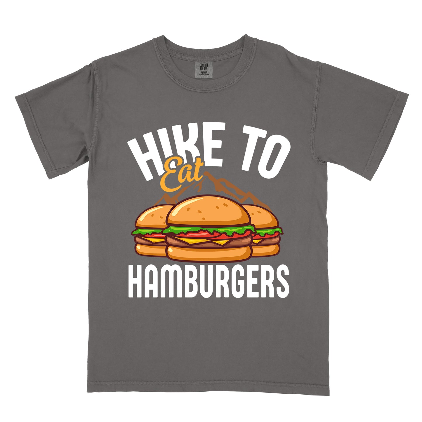 Pepper (Dark Gray) t-shirt with text "HIKE TO Eat HAMBURGERS". "HIKE TO" appears in white arched letters at the top, "Eat" in orange script in the middle, and "HAMBURGERS" in white at the bottom. The center features an illustration of three hamburgers with buns, lettuce, tomato, cheese and beef patties, with brown mountain silhouettes behind them. The design connects hiking activity with food rewards.