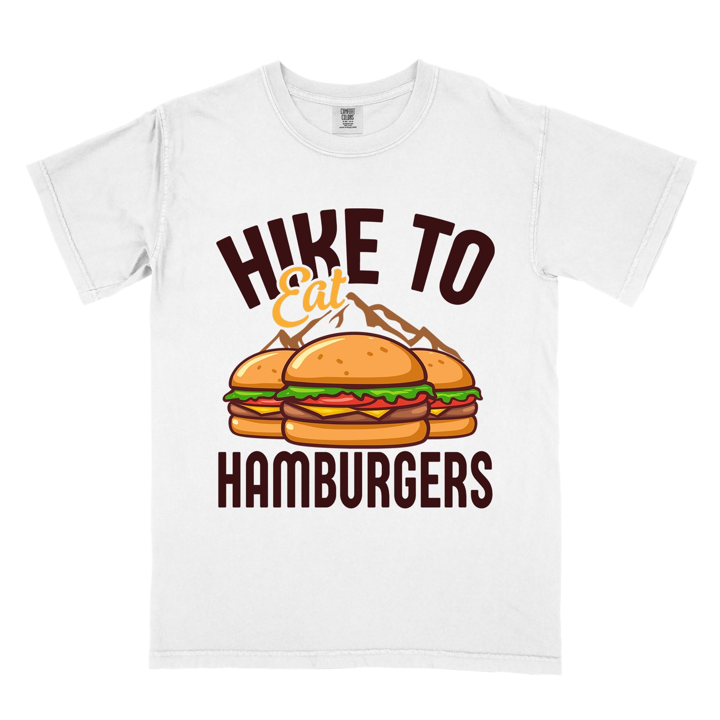 White t-shirt with text "HIKE TO Eat HAMBURGERS". "HIKE TO" appears in deep brown arched letters at the top, "Eat" in orange script in the middle, and "HAMBURGERS" in deep brown at the bottom. The center features an illustration of three hamburgers with buns, lettuce, tomato, cheese and beef patties, with brown mountain silhouettes behind them. The design connects hiking activity with food rewards.
