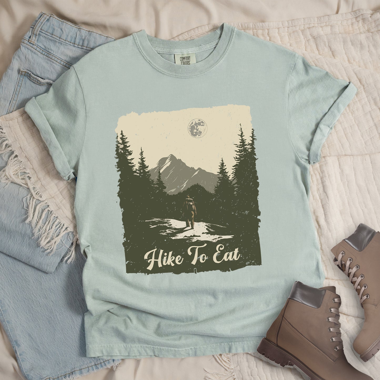 Bay (Light Gray) t-shirt featuring a vintage-style illustration of a lone hiker on a mountain trail. The rectangular design shows pine trees, mountain silhouettes, and a full moon against a cream-colored sky. A solitary hiker with backpack stands on the path facing the mountains. "Hike To Eat" appears in cream script at the bottom of the illustration. The artwork uses muted green, cream, and gray tones in a distressed, rustic style.